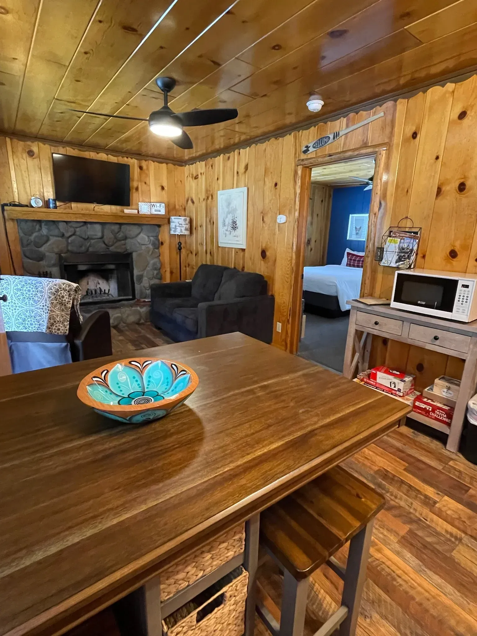 Cozy cabin interior: wooden walls, fireplace, kitchen island with turquoise bowl, doorway to bedroom.