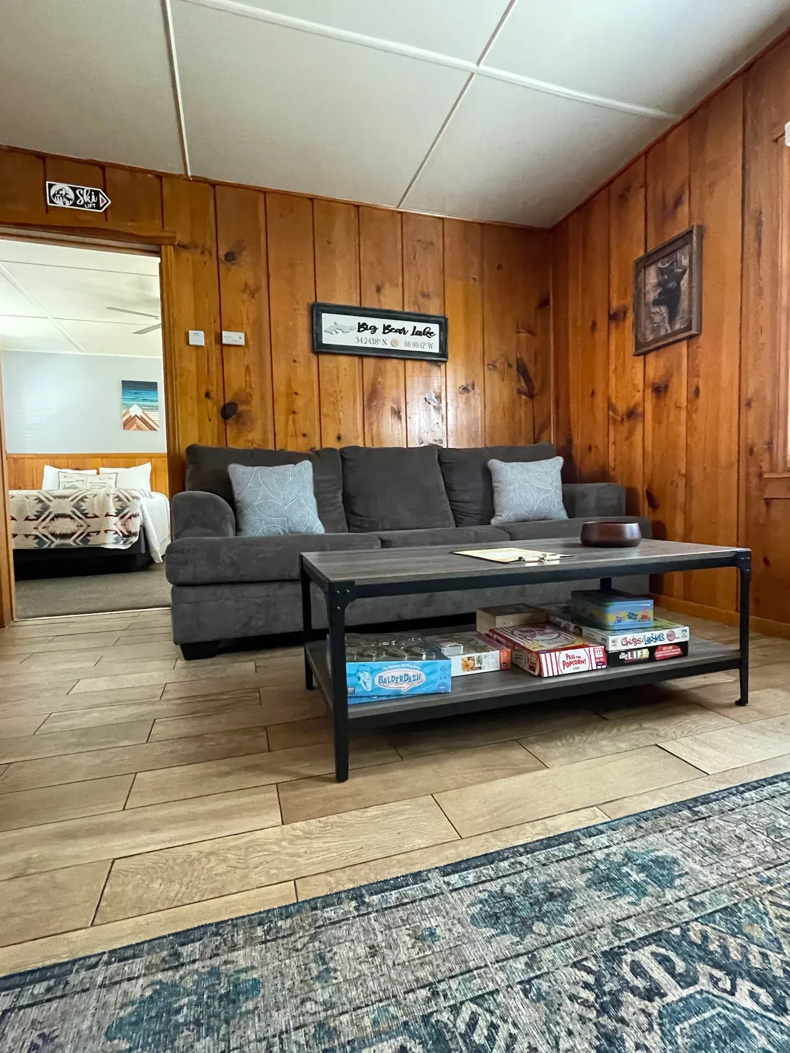 Cozy living room with brown wood paneling, gray sofa, coffee table with games, and an open doorway to a bedroom.