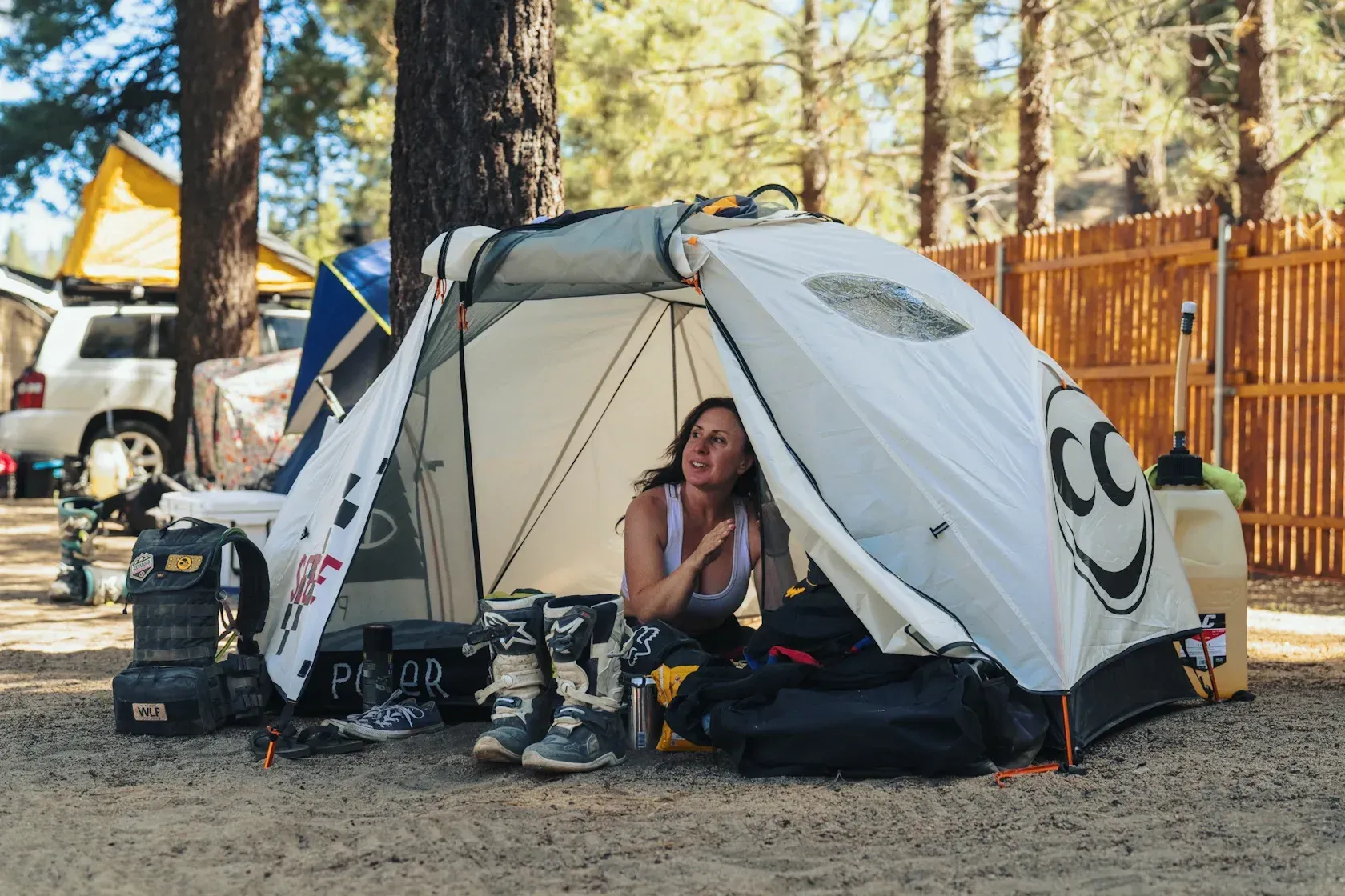 Woman inside a tent, smiling, surrounded by camping gear. Sunny outdoor setting.