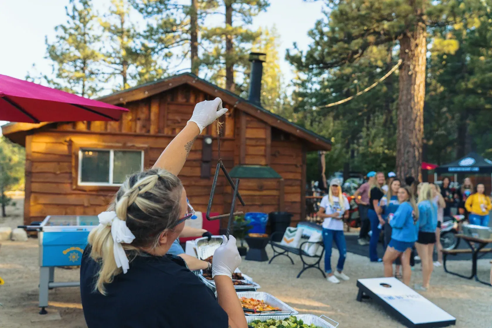 Woman serving food outside a wooden cabin; a group of people is nearby playing games.