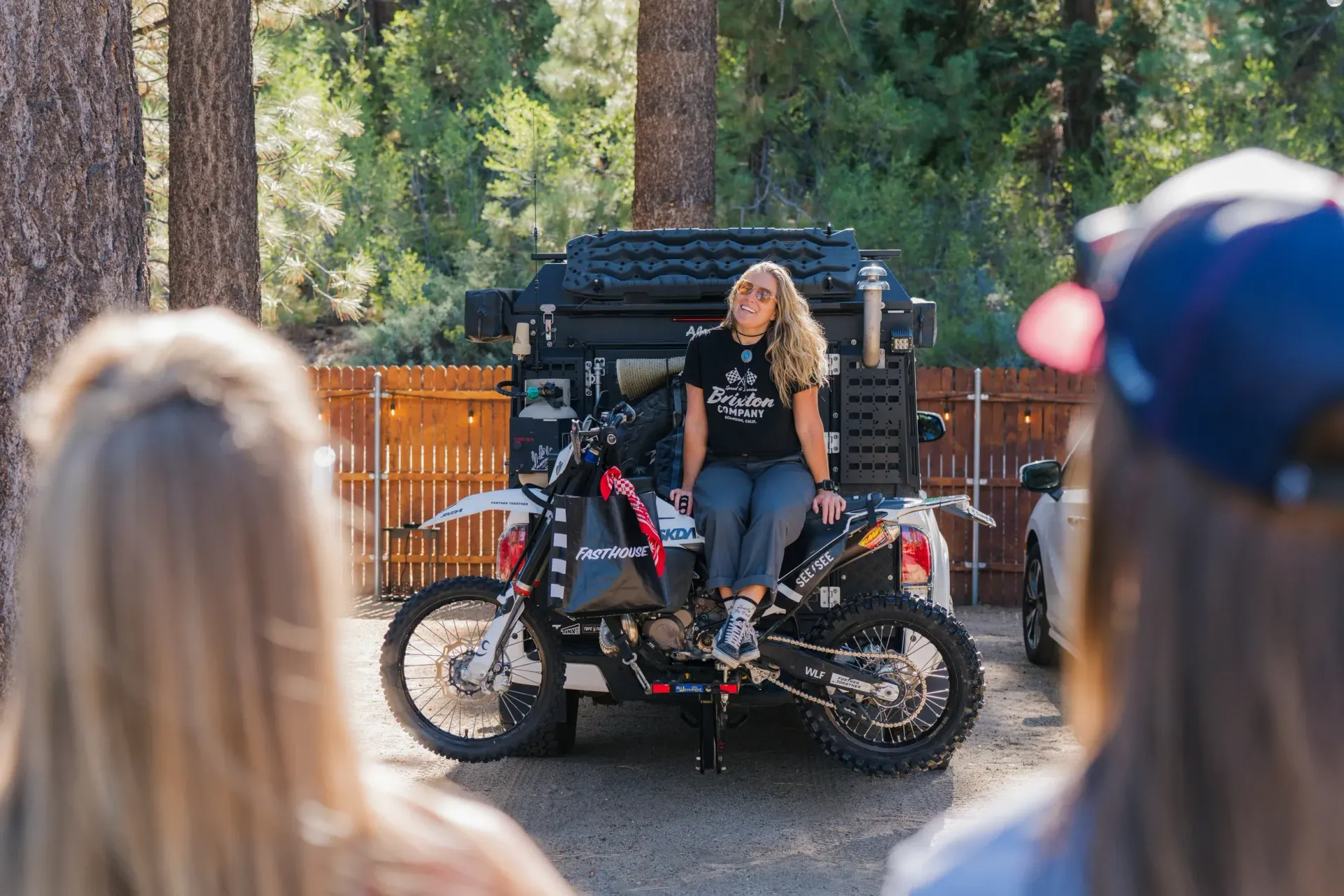 Woman on a modified motorcycle smiles, sitting in front of two friends. Motorcycle is loaded with gear, surrounded by trees.