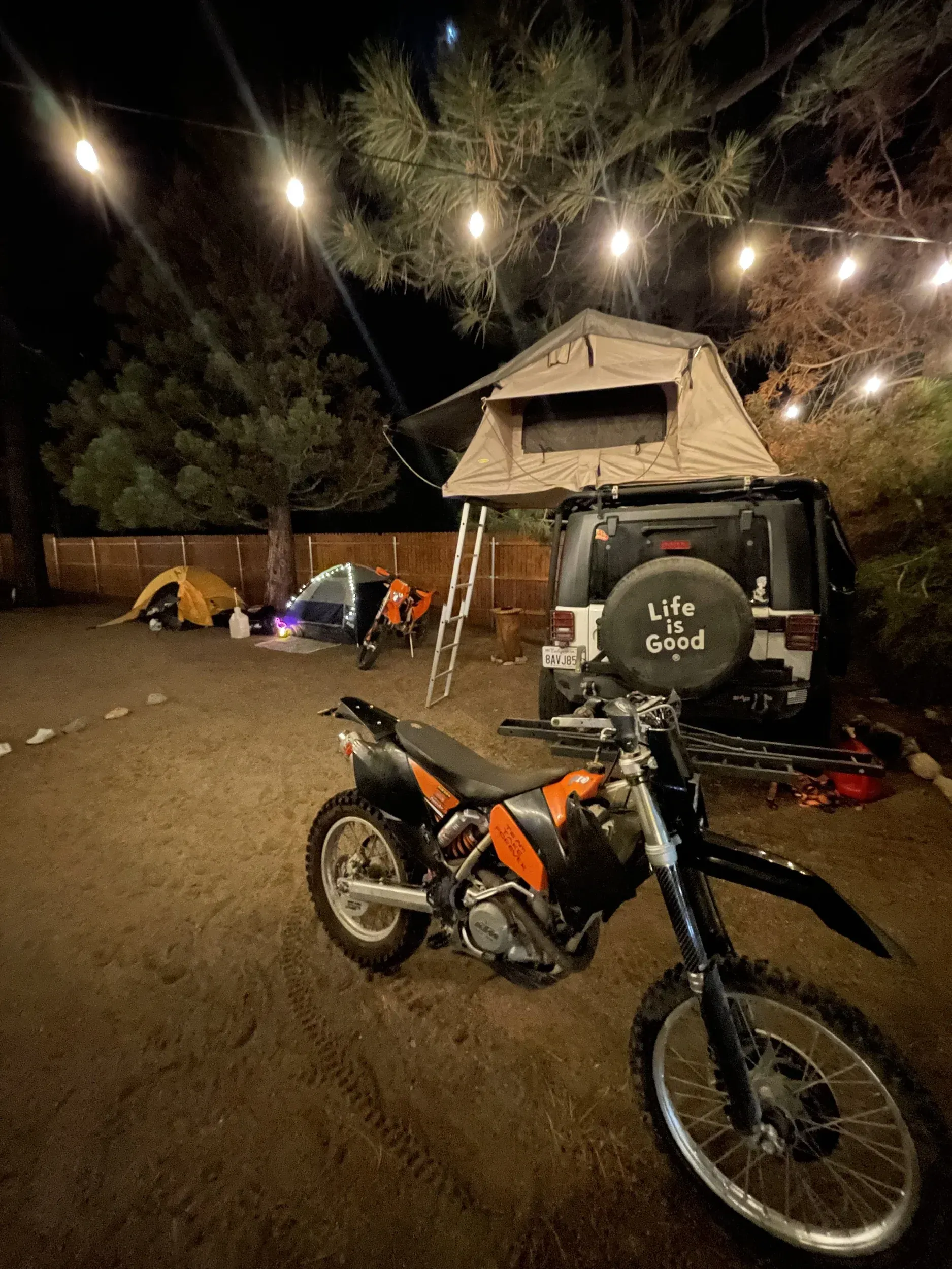 Motorcycle parked near a Jeep with a rooftop tent at a campsite at night, string lights overhead.