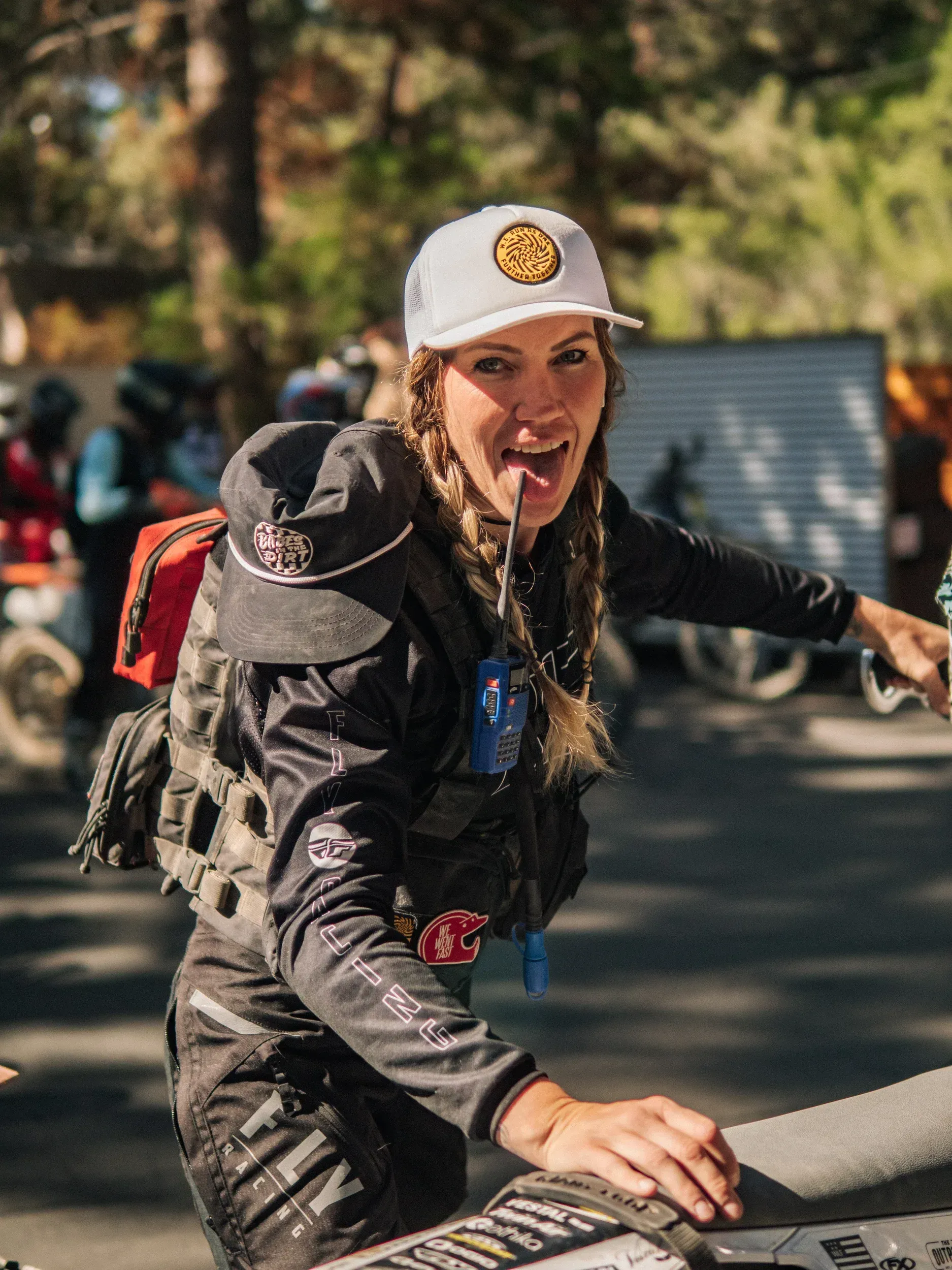 Woman wearing a white cap and black jacket, sticks out tongue, smiles. Has a small backpack, and is in an outdoor setting.