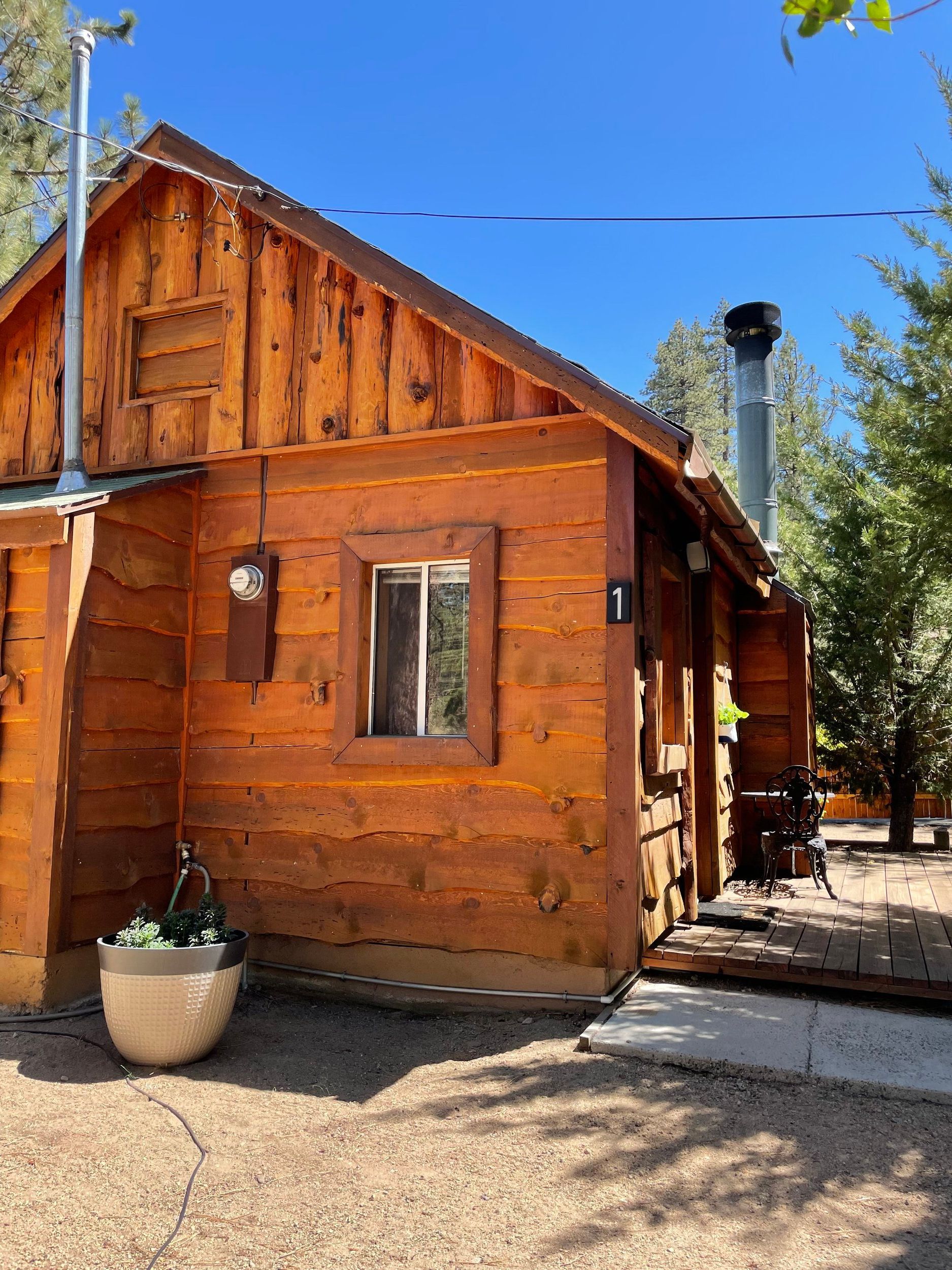 Wooden cabin with a window, chimney, and hanging flower pot, under a blue sky.