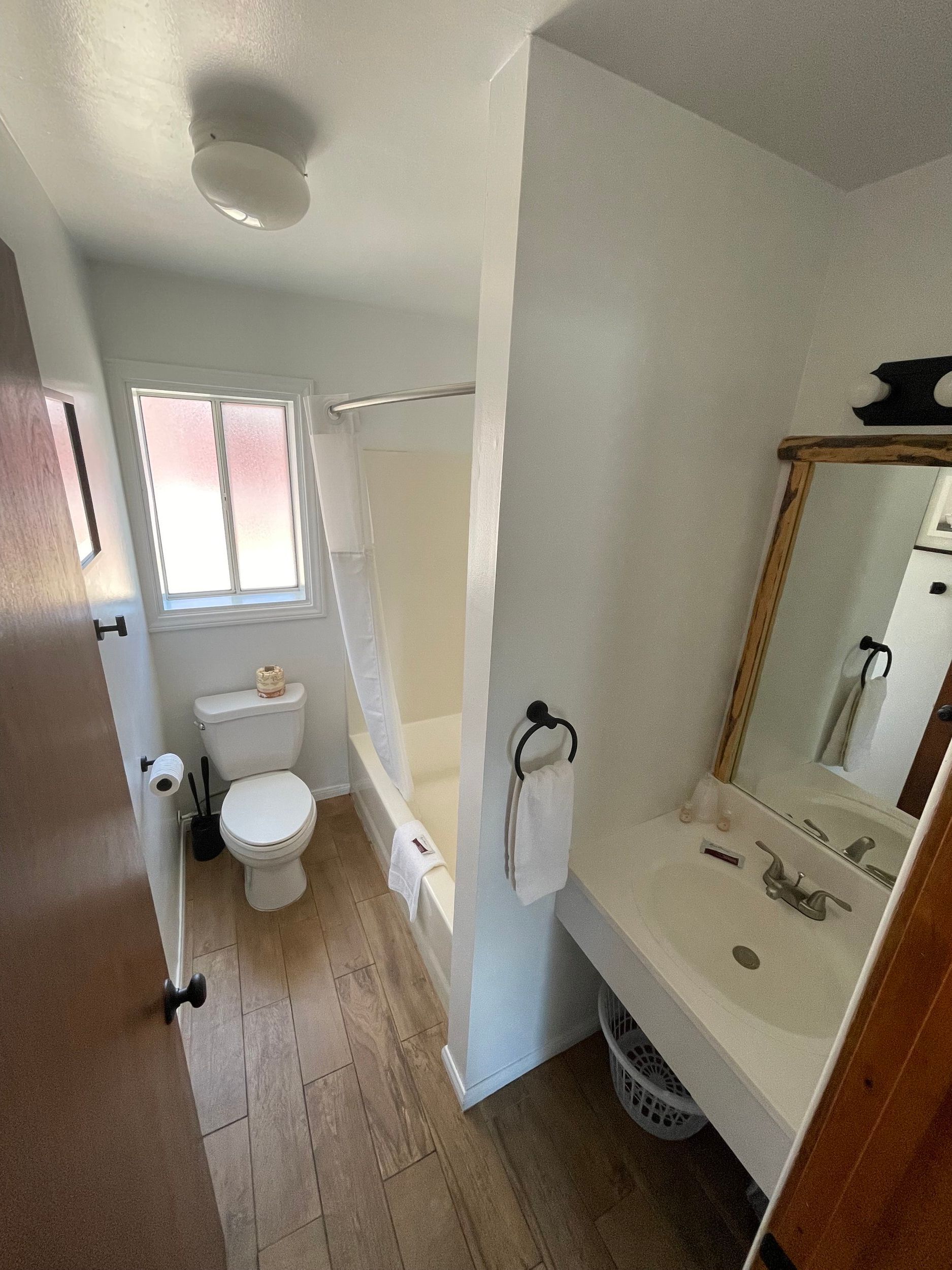 Bathroom with toilet, tub/shower, sink, window, and wood-look flooring.
