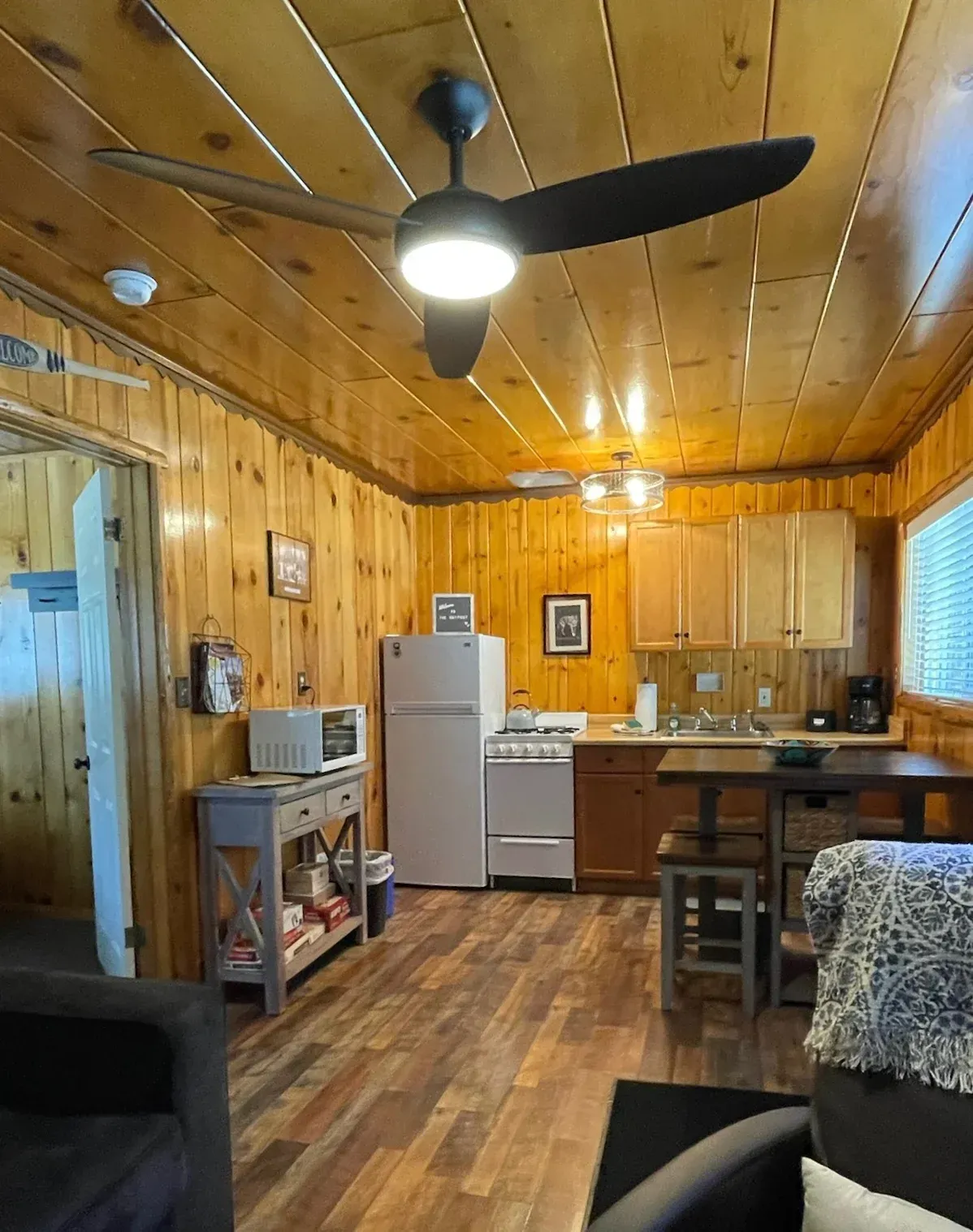 Small cabin interior with wood paneling, kitchen, furniture, and a ceiling fan.
