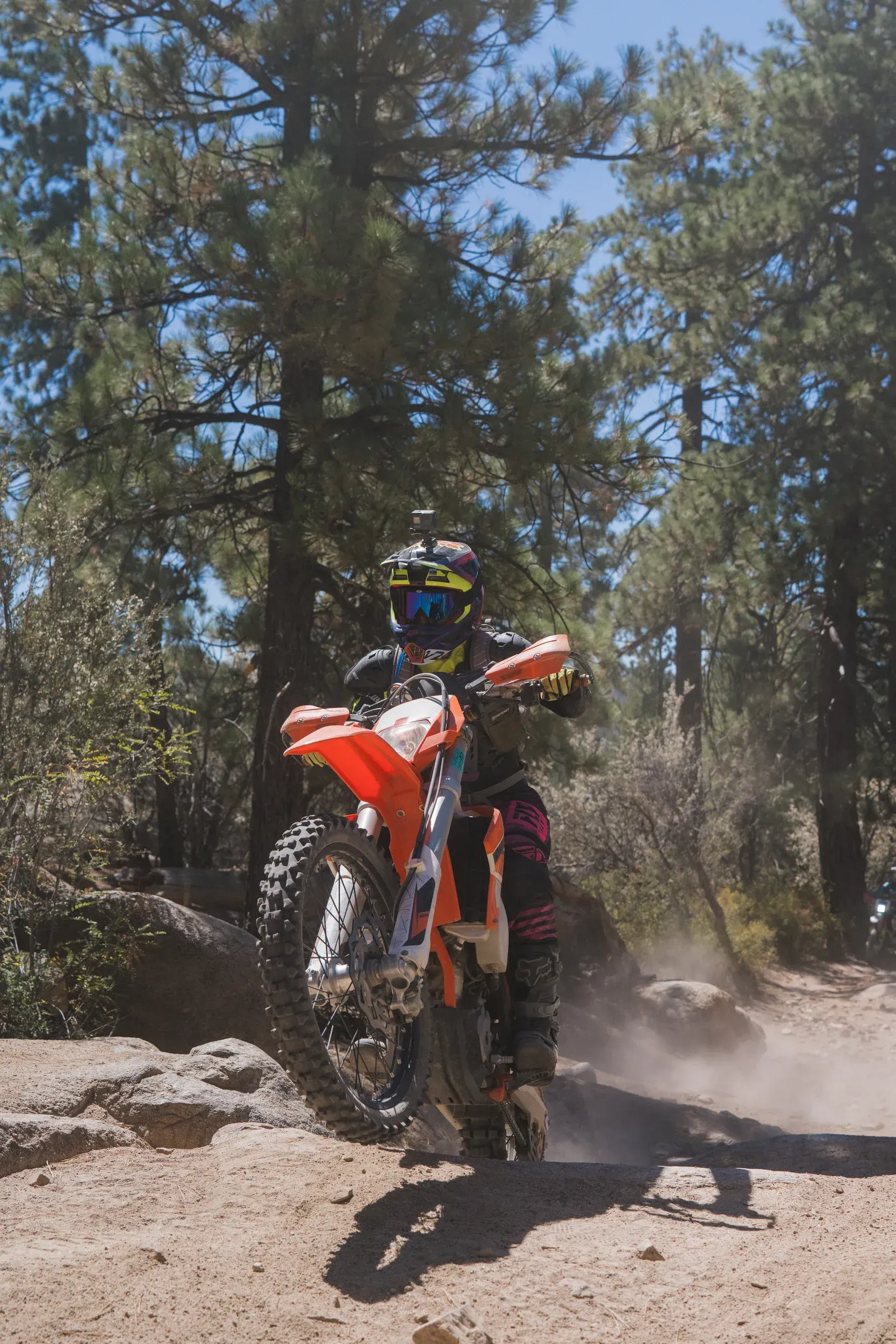 Dirt bike rider on a rocky dirt path, kicking up dust. Orange bike, rider wearing protective gear, trees in background.