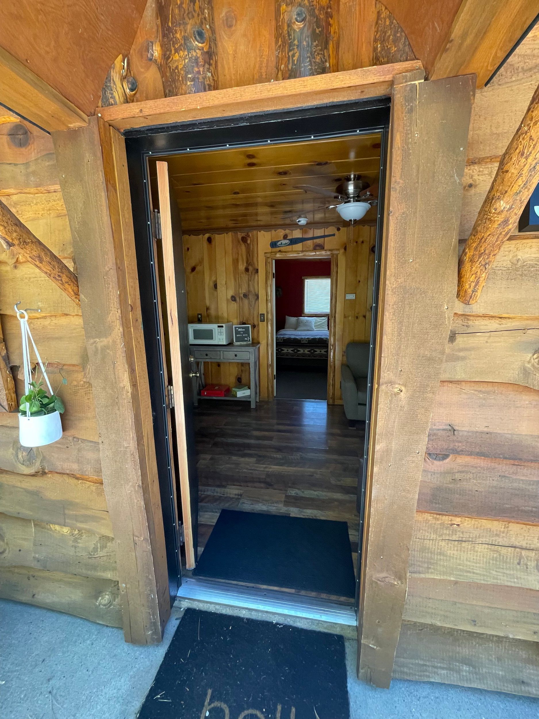Entryway of a rustic cabin, showing the interior. Doorway framed by wood. A welcome mat, microwave, and bed are visible.
