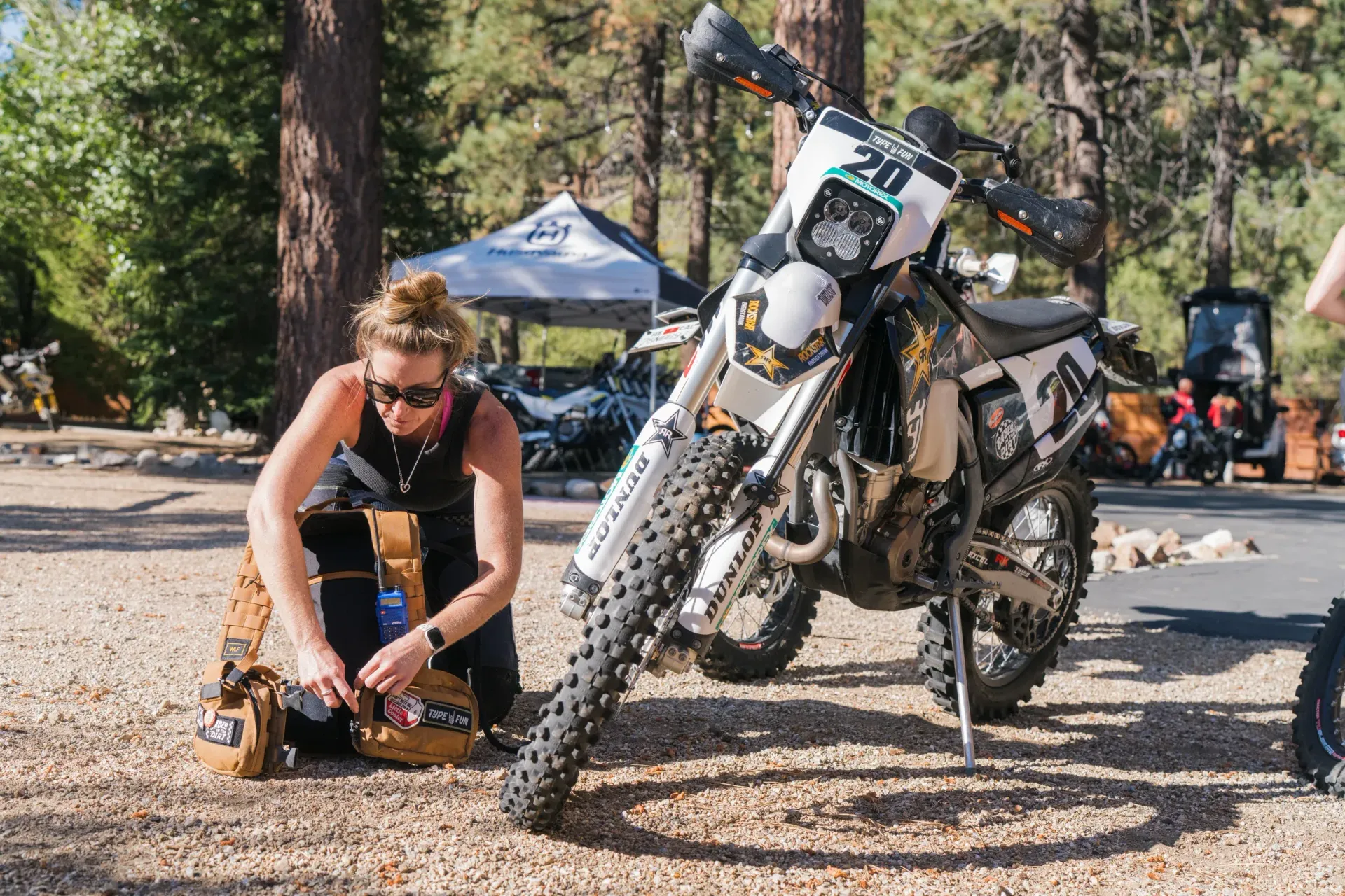 Woman kneeling, packing bag beside a parked motorcycle on gravel, trees in background.