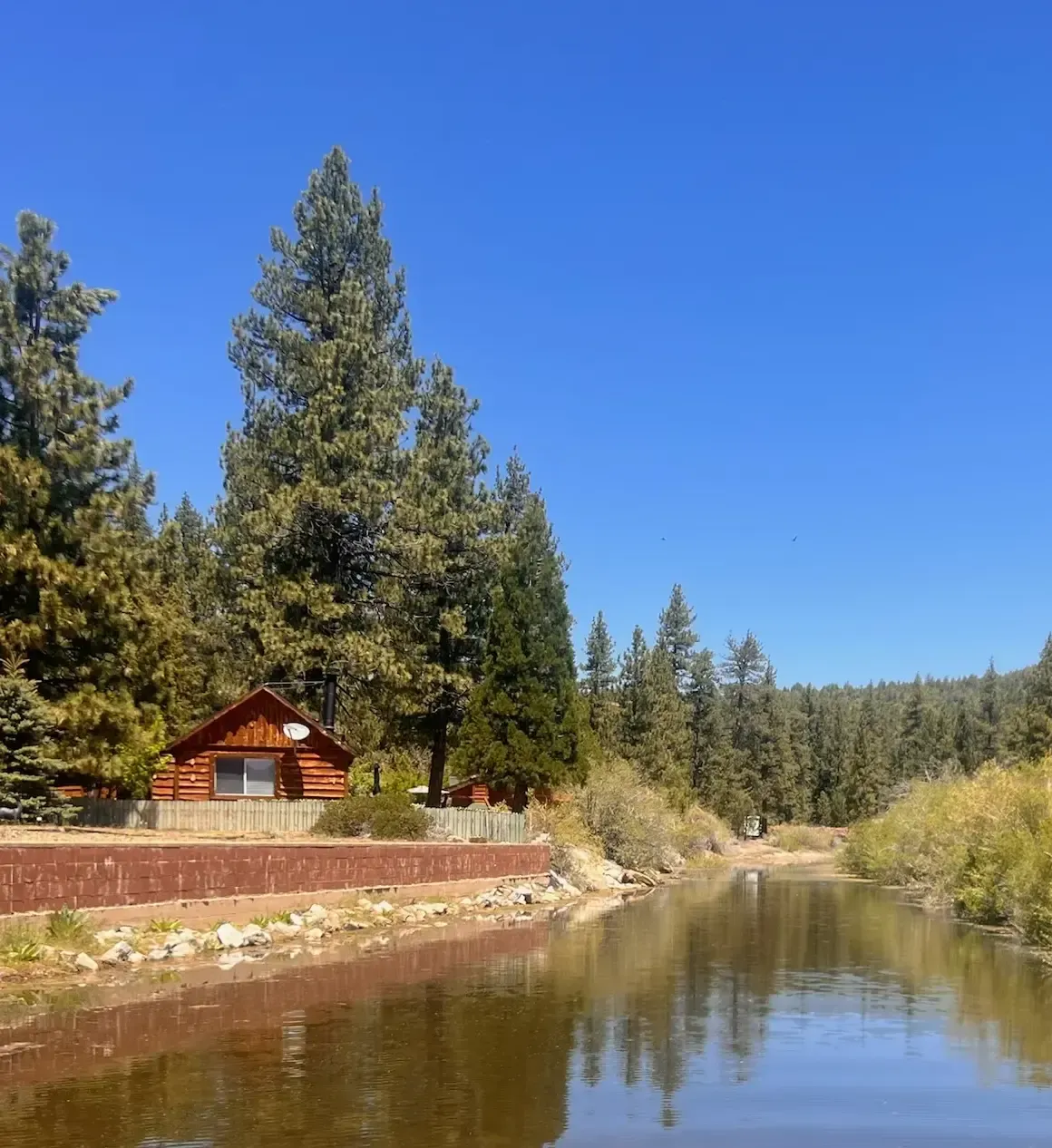 Cabin by a calm river with trees under a bright blue sky.