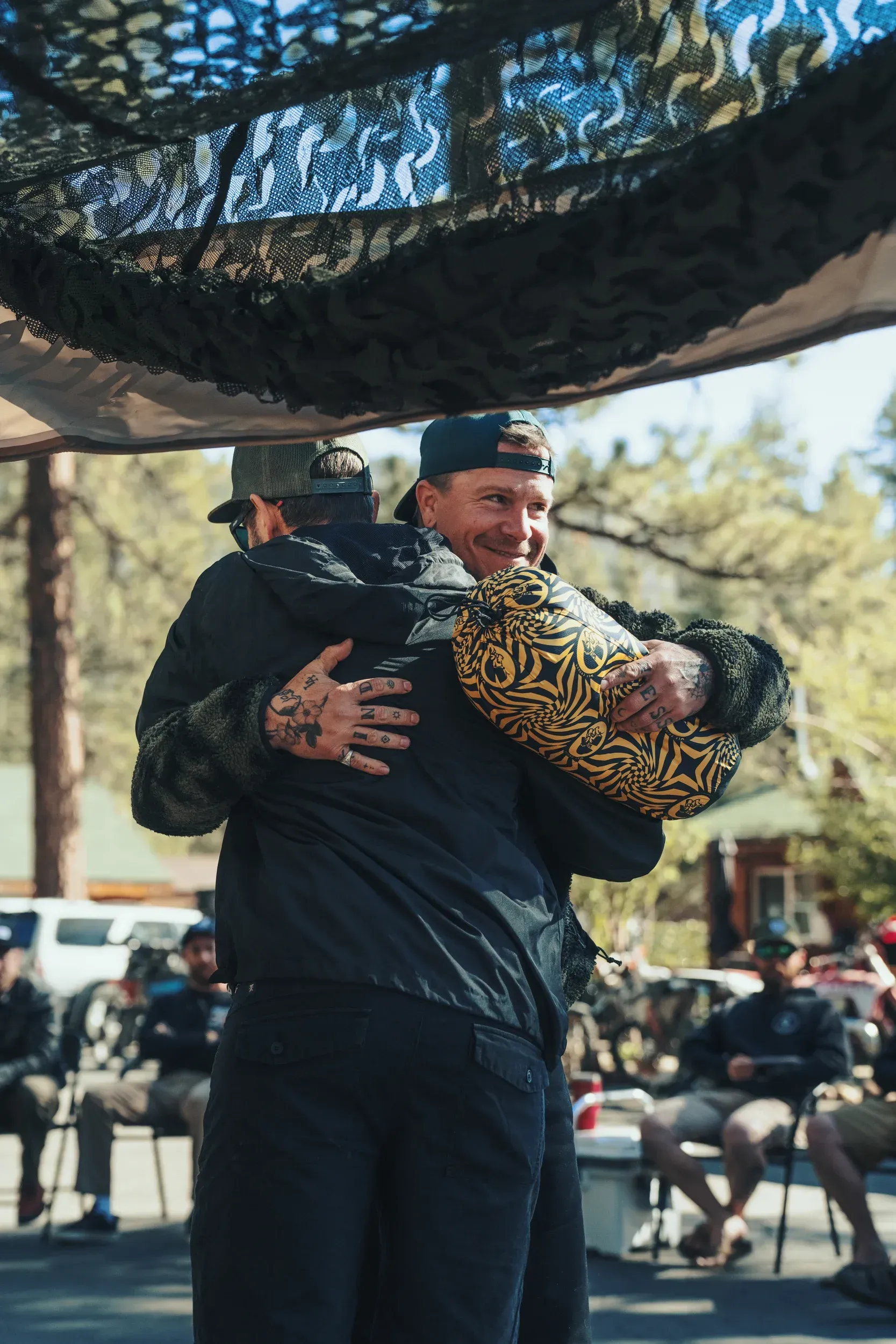 Two men embracing under camouflage netting outdoors. One is wearing a hat. Others sit nearby.