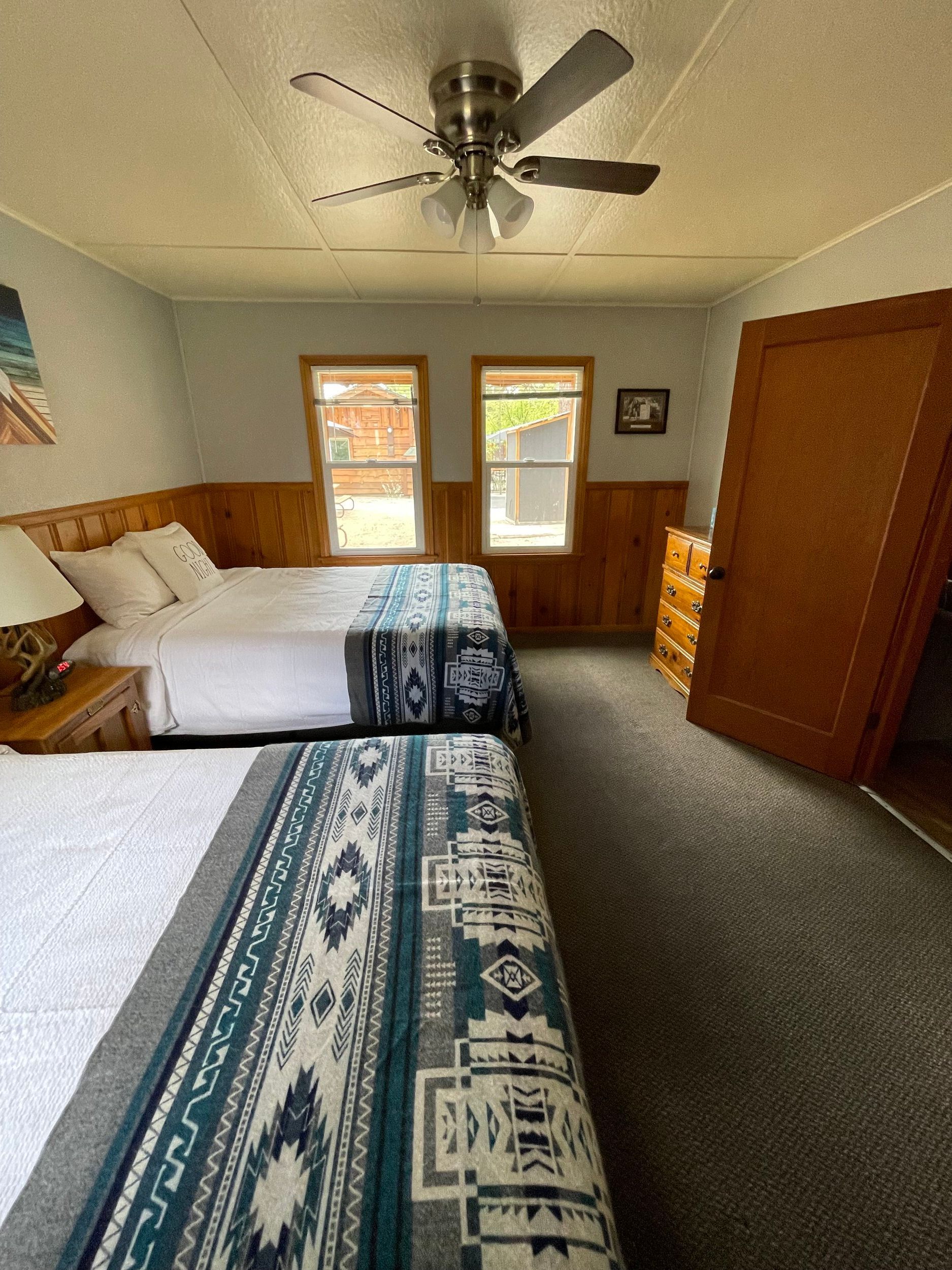 Bedroom with two double beds, patterned blankets, wood paneling, windows, ceiling fan, and a dresser.