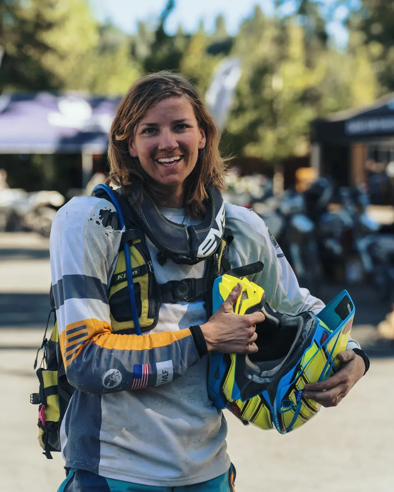 Person in motorcycle gear smiles, holding a helmet. Outdoors, with a forest background.