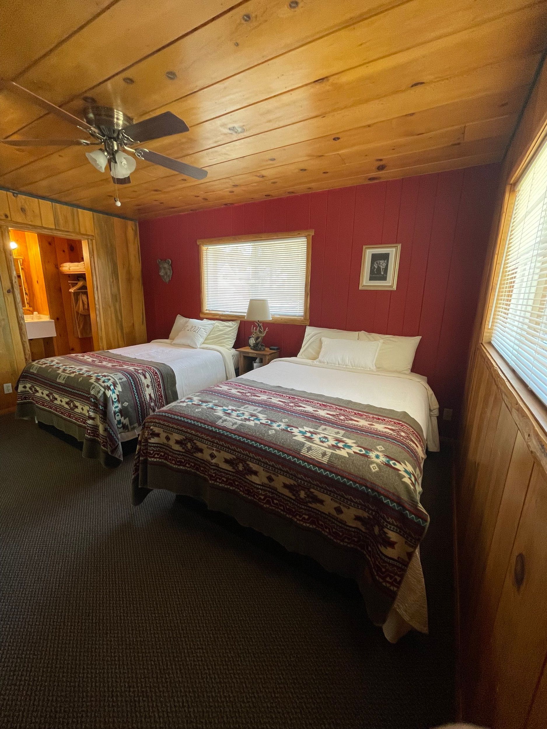 Bedroom with two double beds, red wall, patterned blankets, wood ceiling and walls, and bathroom entrance.