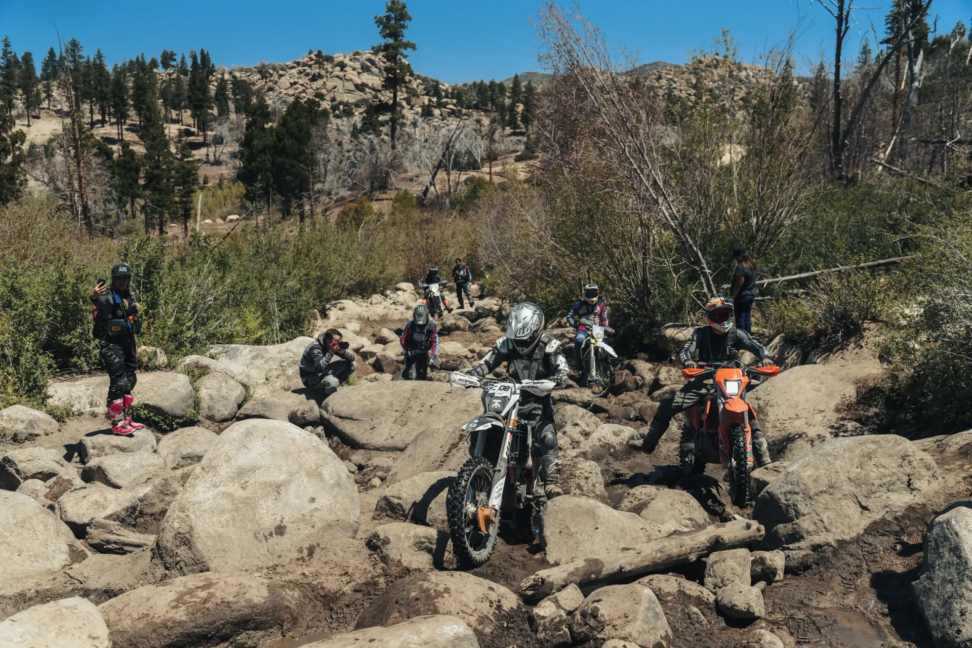 Dirt bikers navigating a rocky, muddy trail in a mountainous setting, under a bright blue sky.