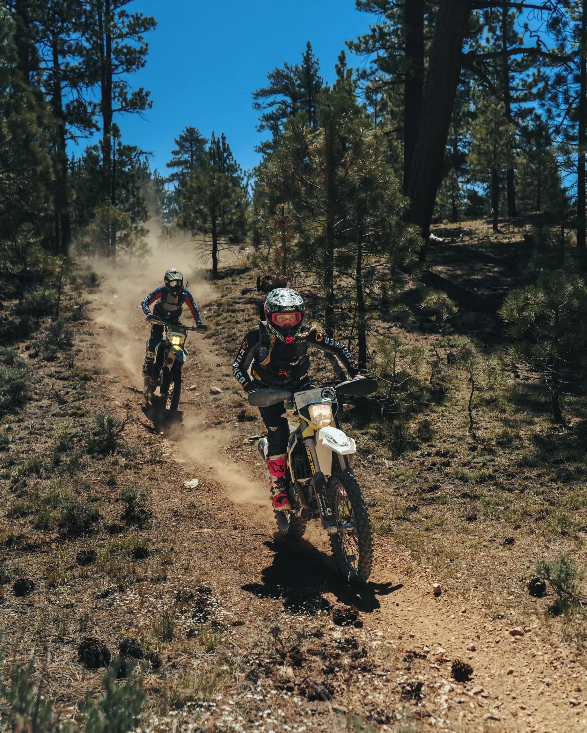 Two dirt bikes riding on a dusty trail through a forest under a bright blue sky.