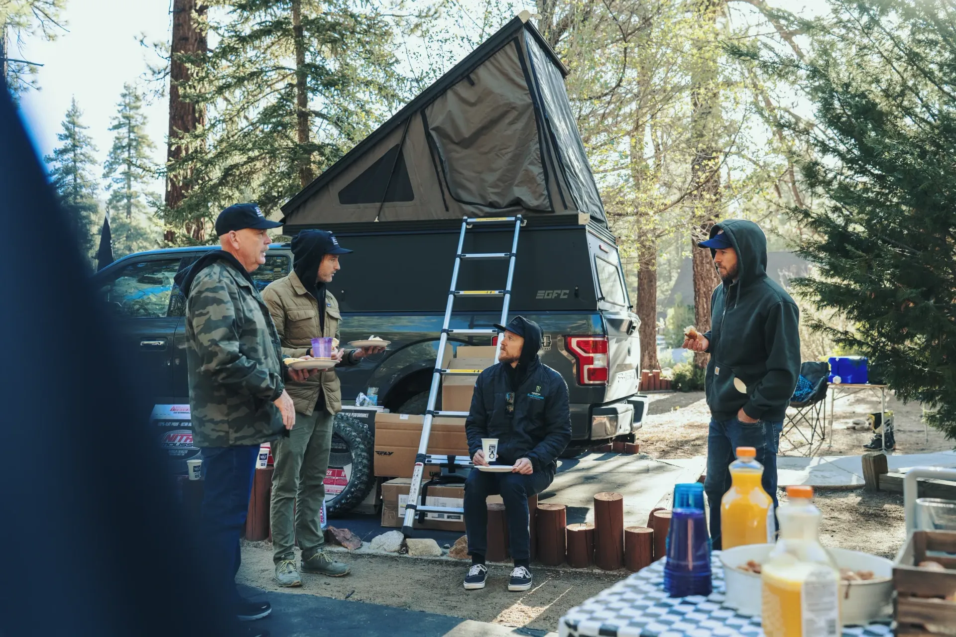 Group of people gathered outdoors by a truck with a rooftop tent. They are talking and eating.