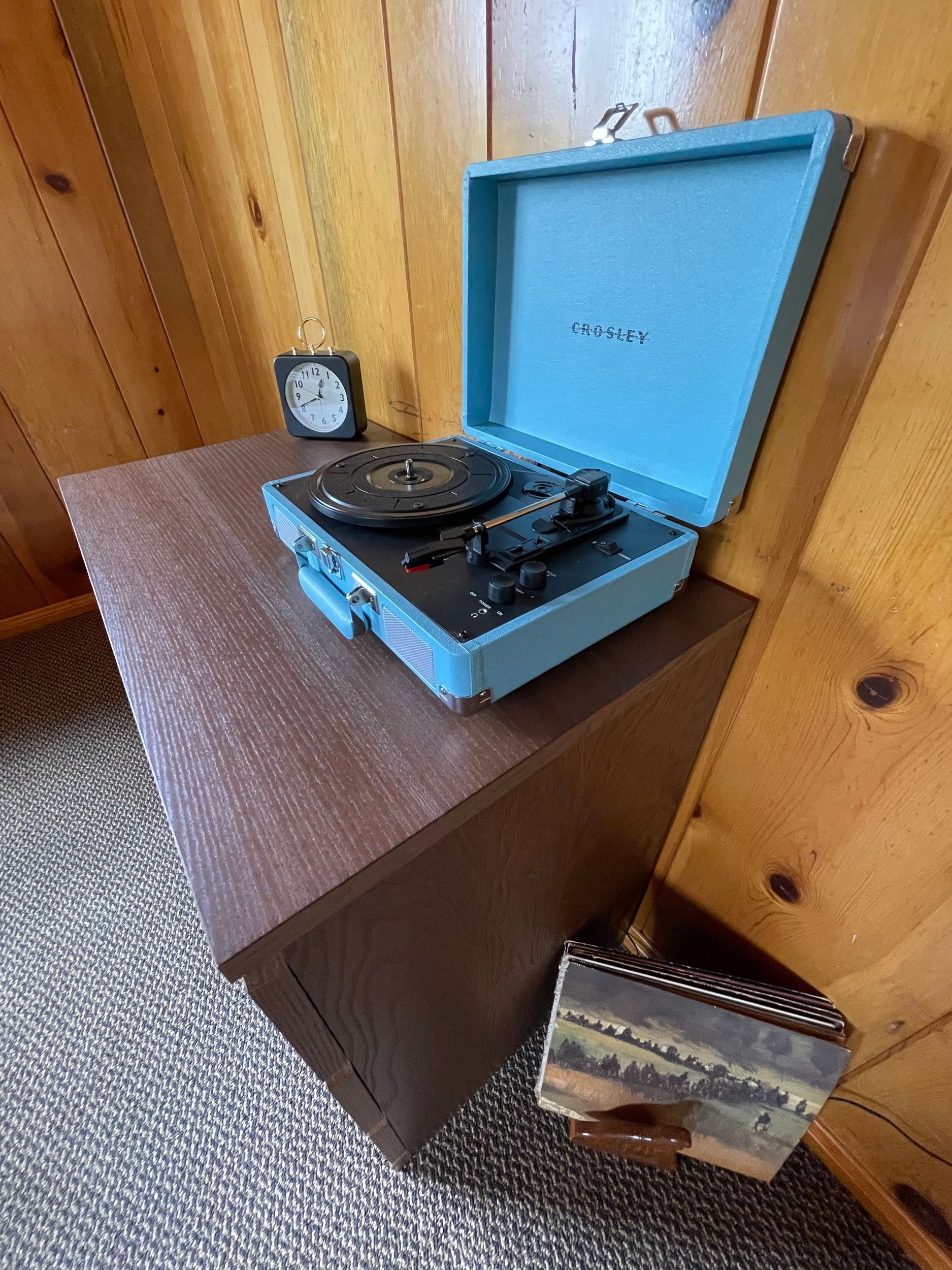 Blue record player on a brown side table next to a clock and a small decorative box, wooden wall background.