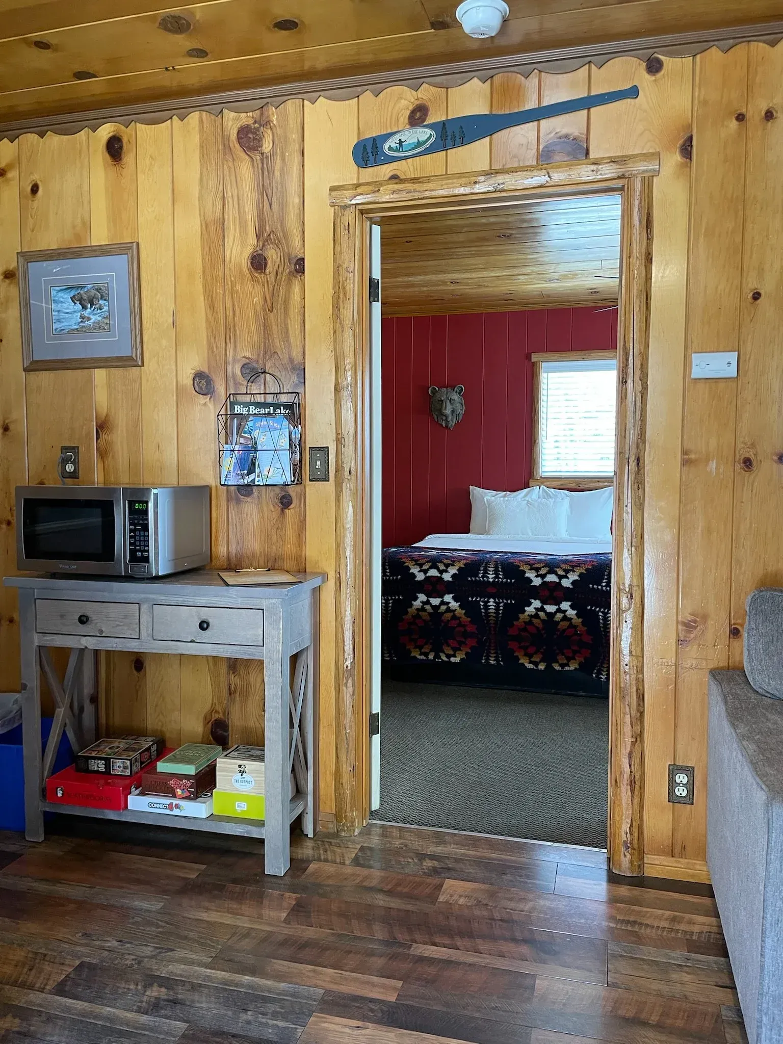 Cabin interior with wood paneling, microwave, table, doorway to a bedroom with bed and red wall.