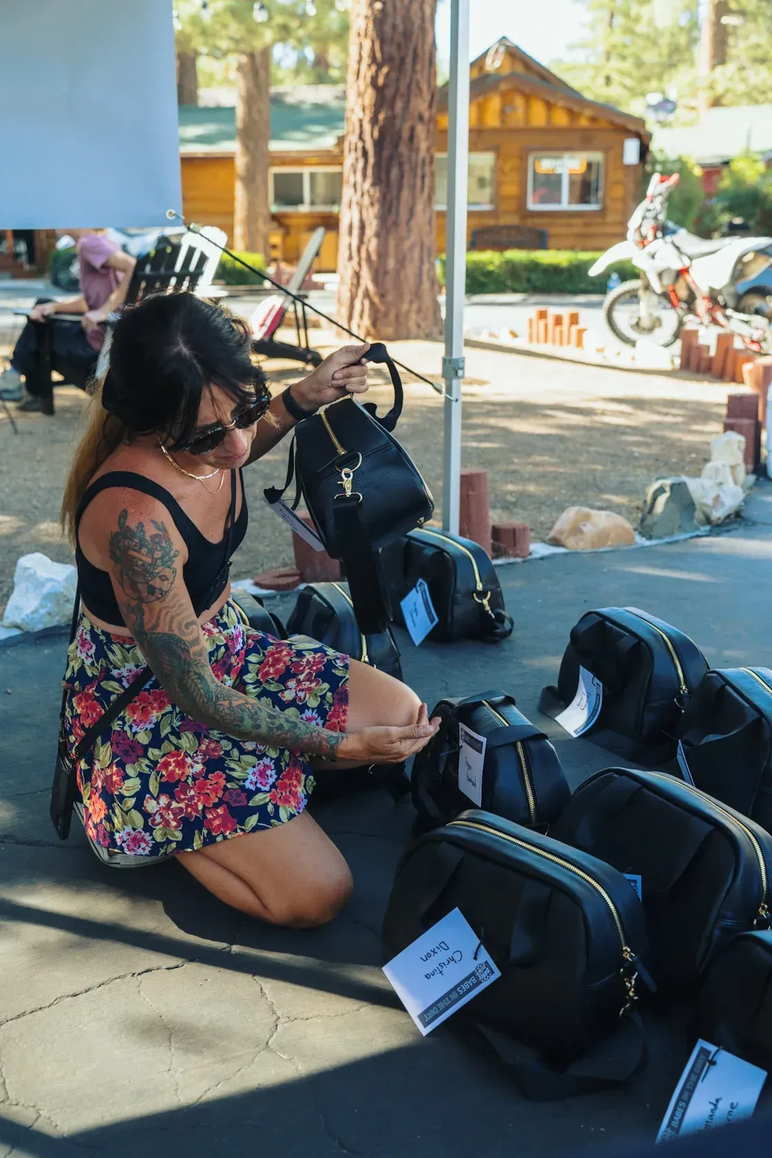 Woman kneeling, arranging black backpacks with tags outdoors.