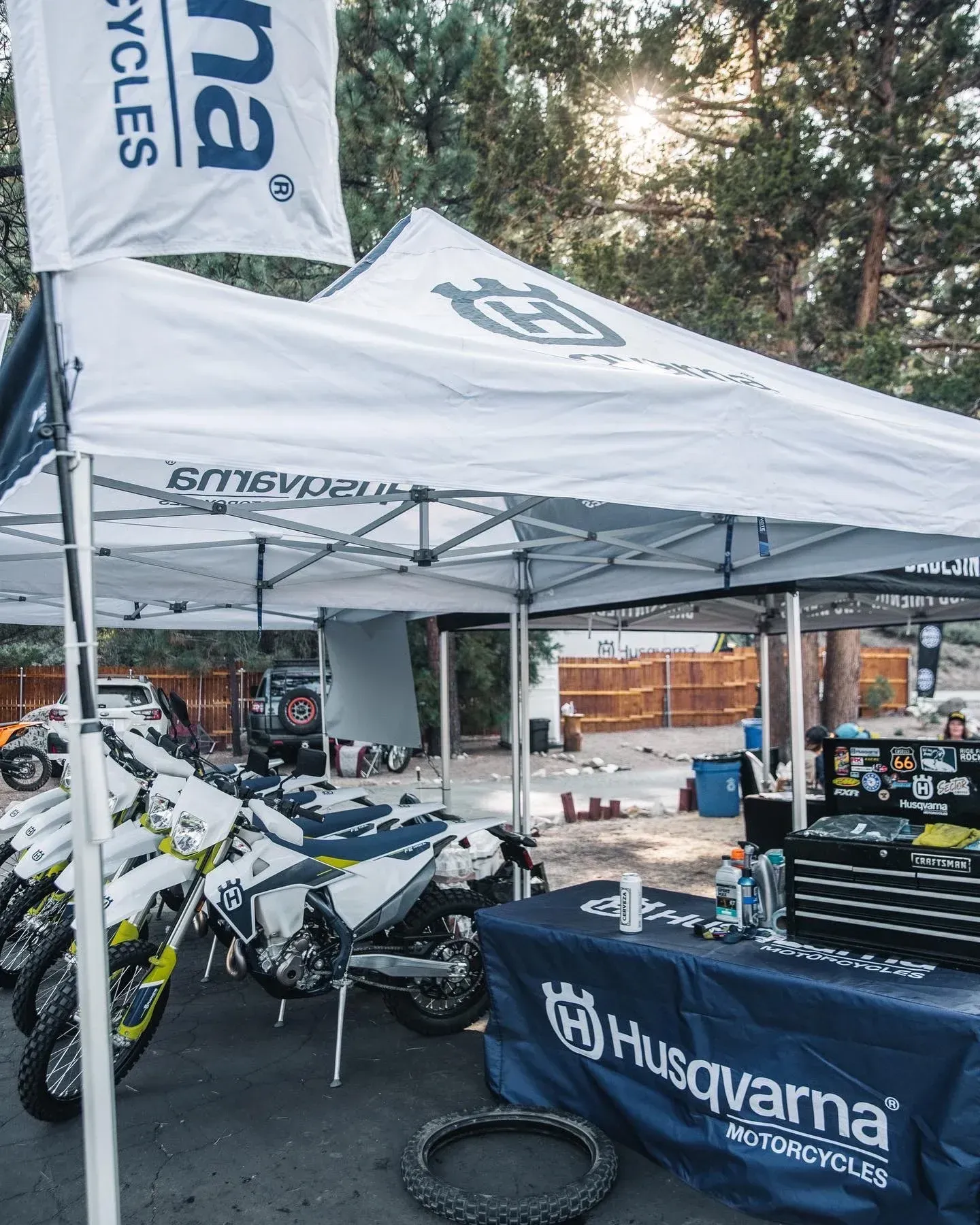 Husqvarna motorcycles displayed under a branded tent at an outdoor event. Workshop tools, spare tire, and blue banner present.