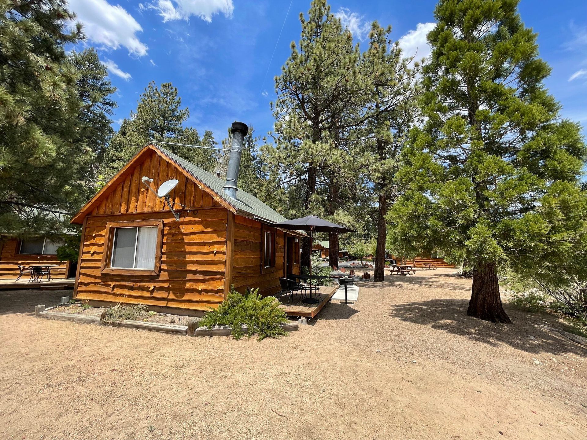 Log cabin with a chimney, satellite dish, and patio, surrounded by trees and gravel.