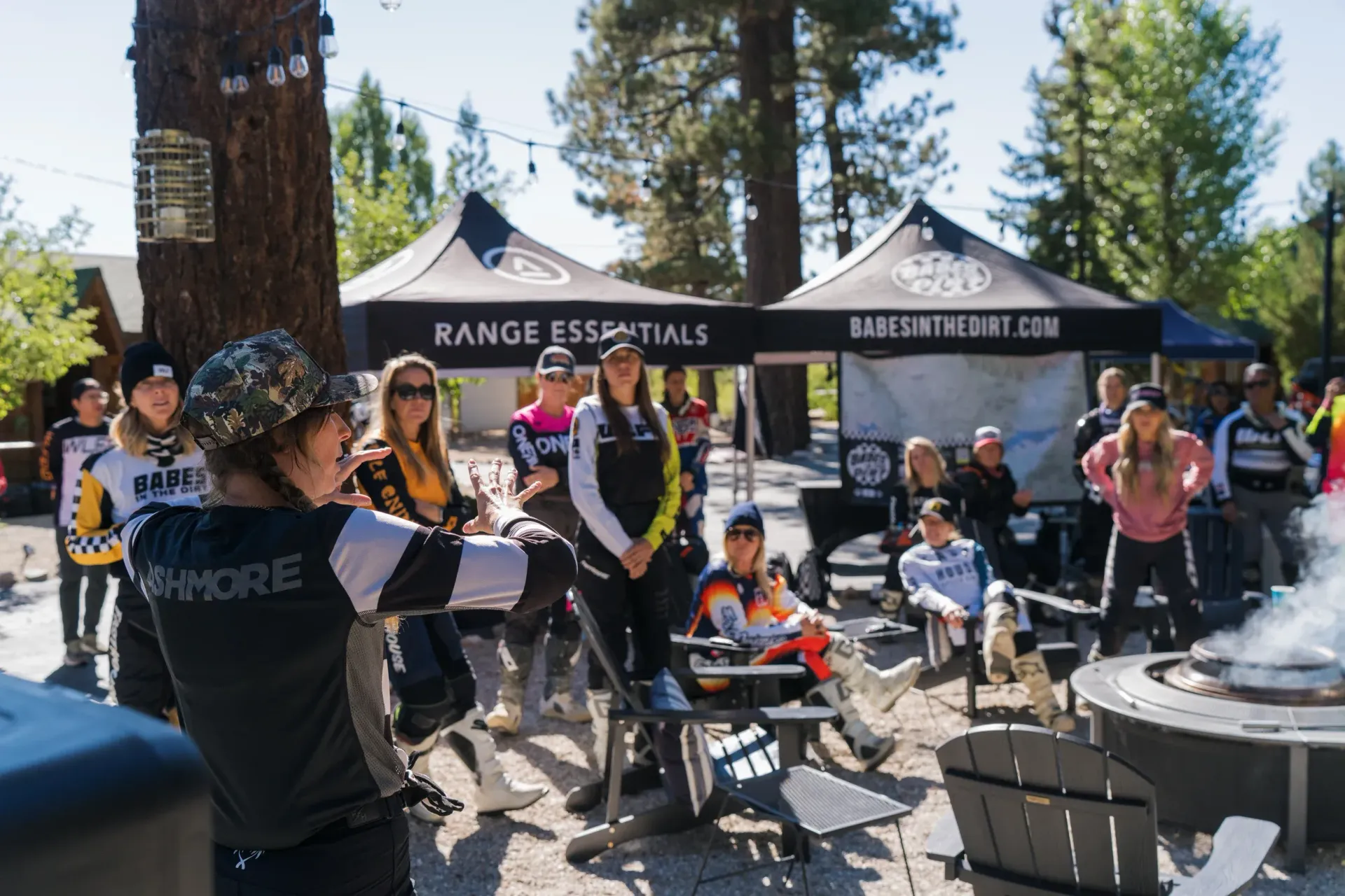 Group outdoors near fire pit, listening. Tents, trees, and casual setting.