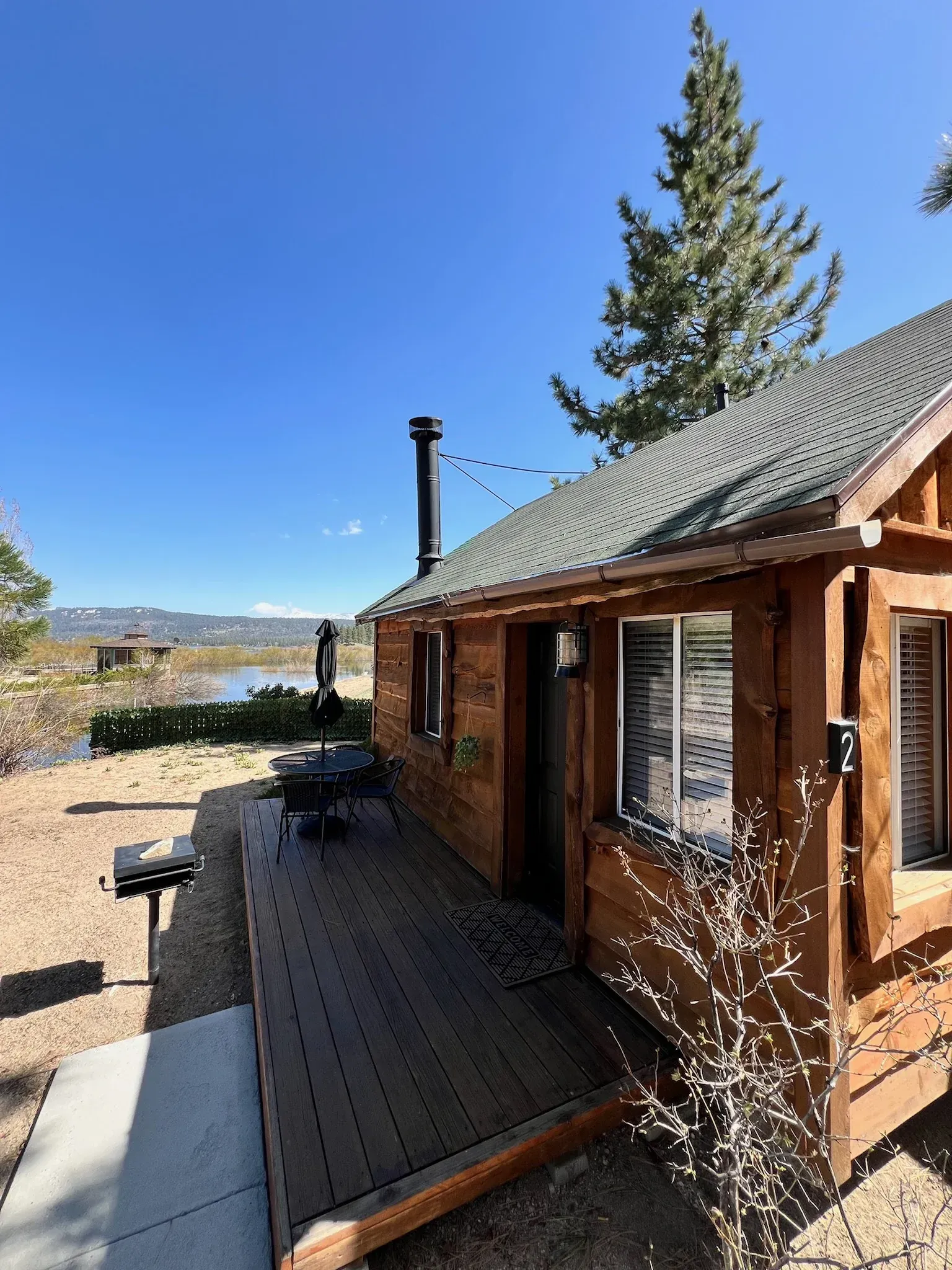 Wooden cabin with deck, overlooking a lake. Sunny day with blue sky and trees.