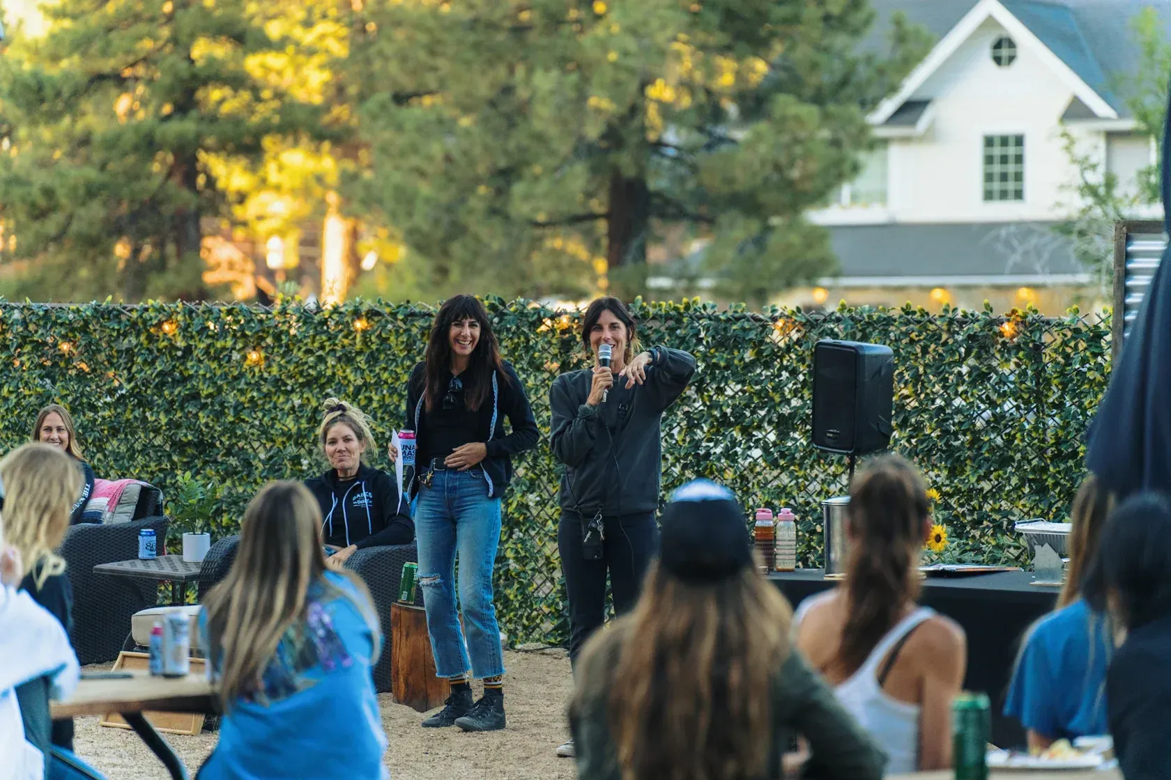 Two people stand on a stage, speaking to an outdoor audience. The stage is in a backyard.
