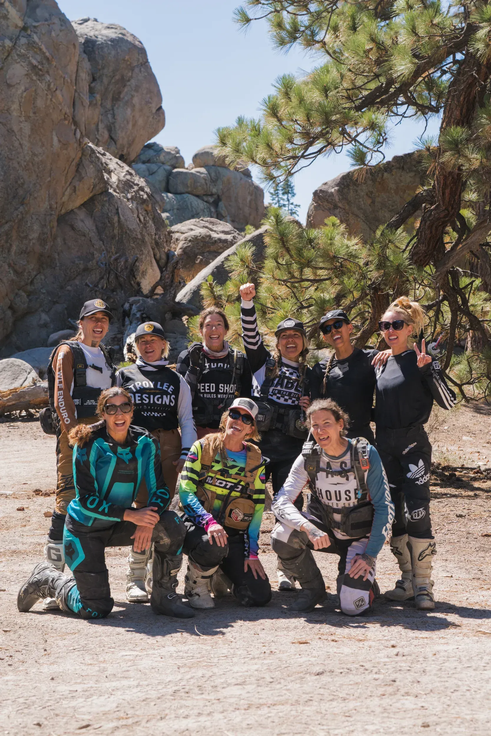 Group of people in motocross gear pose on a dirt path in a rocky, wooded area.