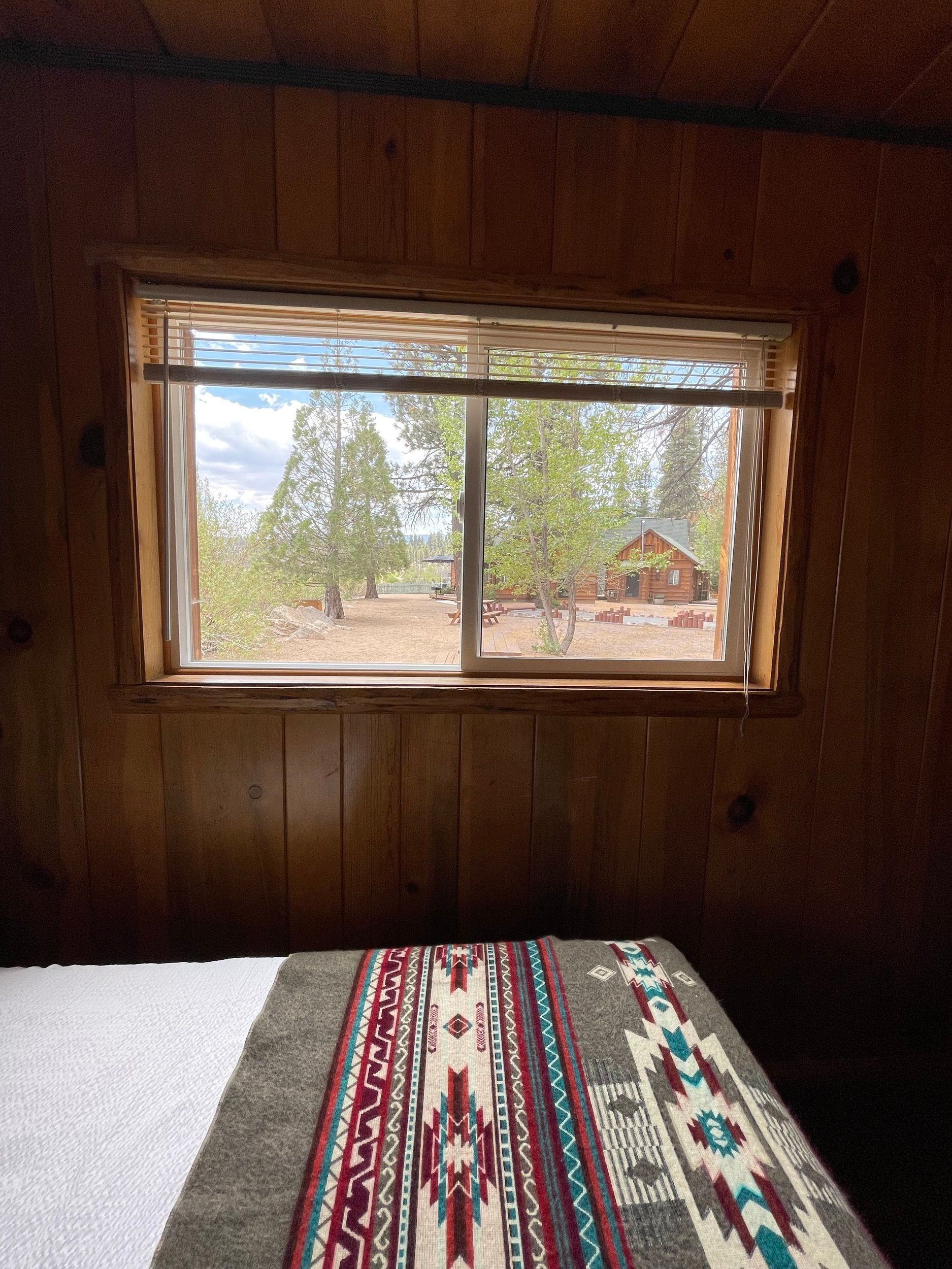 Bedroom window with view of trees and a distant cabin. A patterned blanket lies on the bed.