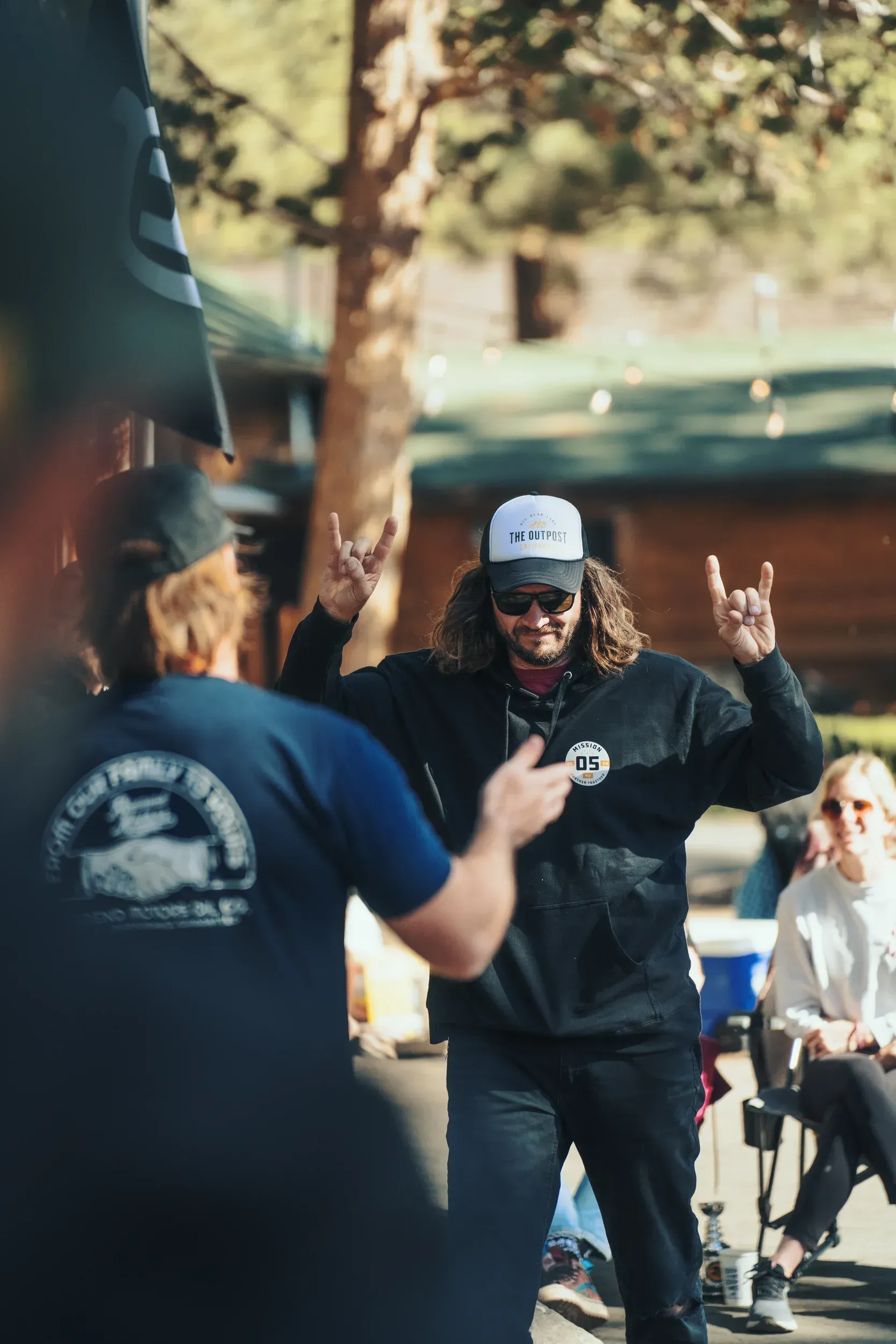 Man in white hat and black hoodie makes rock-on gesture in front of another man.