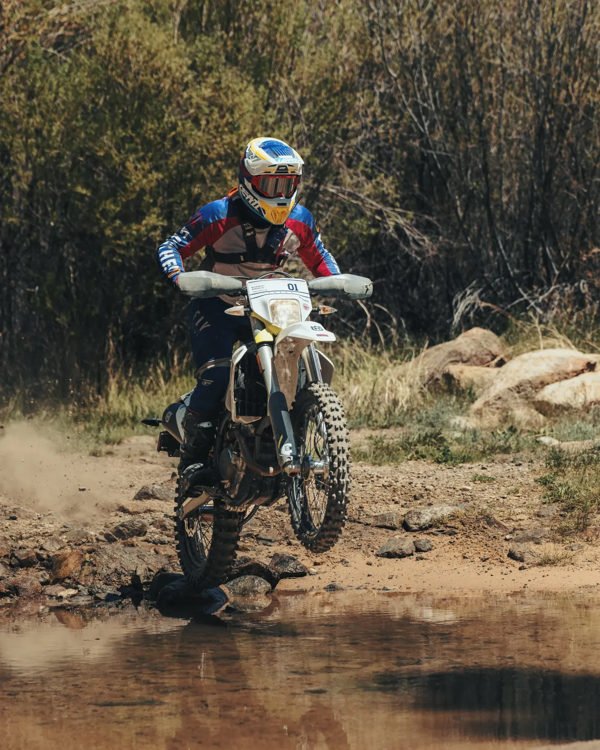 Dirt bike rider in blue and red gear, lifting front wheel over water, dirt road.
