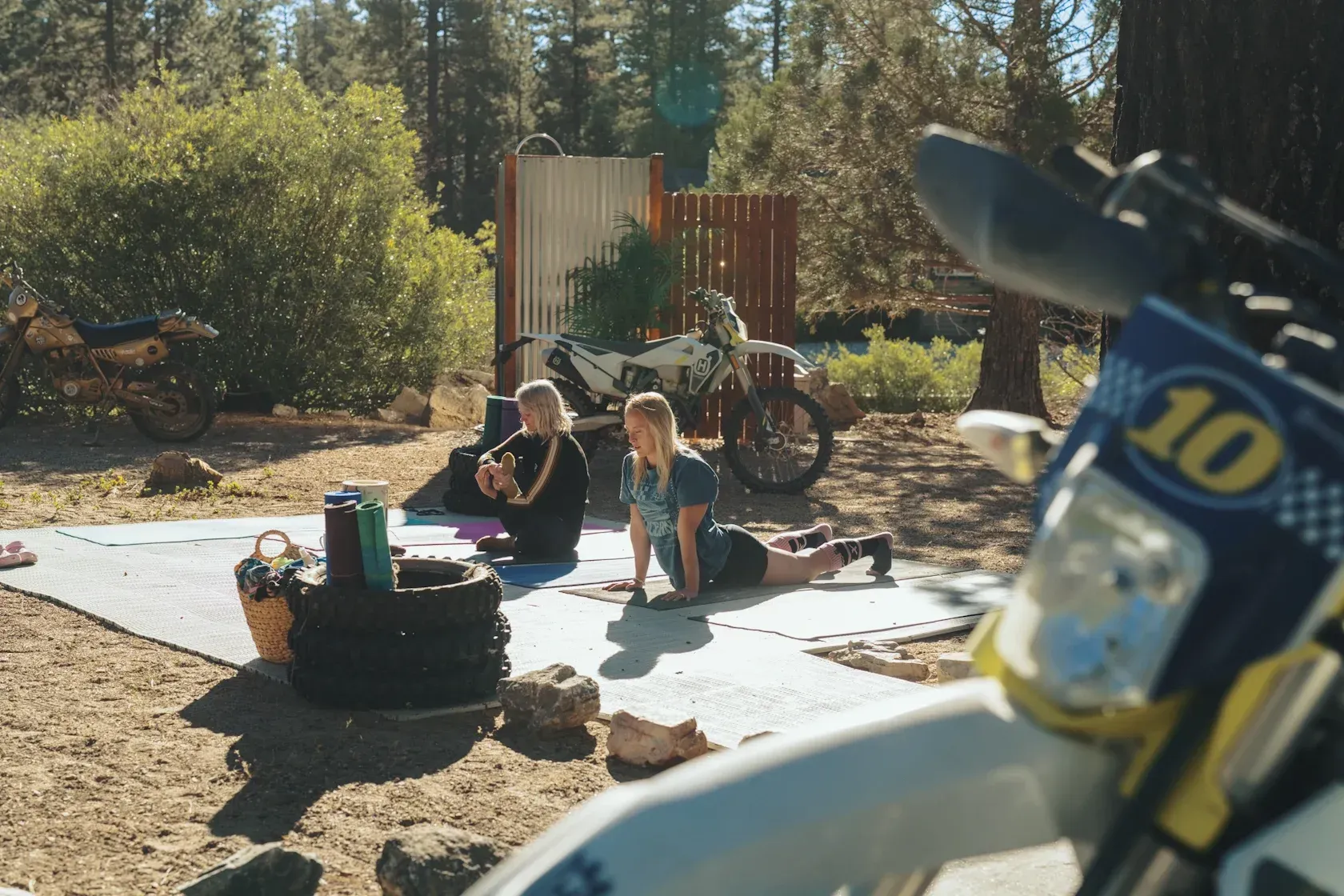 Two people doing yoga on a mat in a wooded area near motorcycles.
