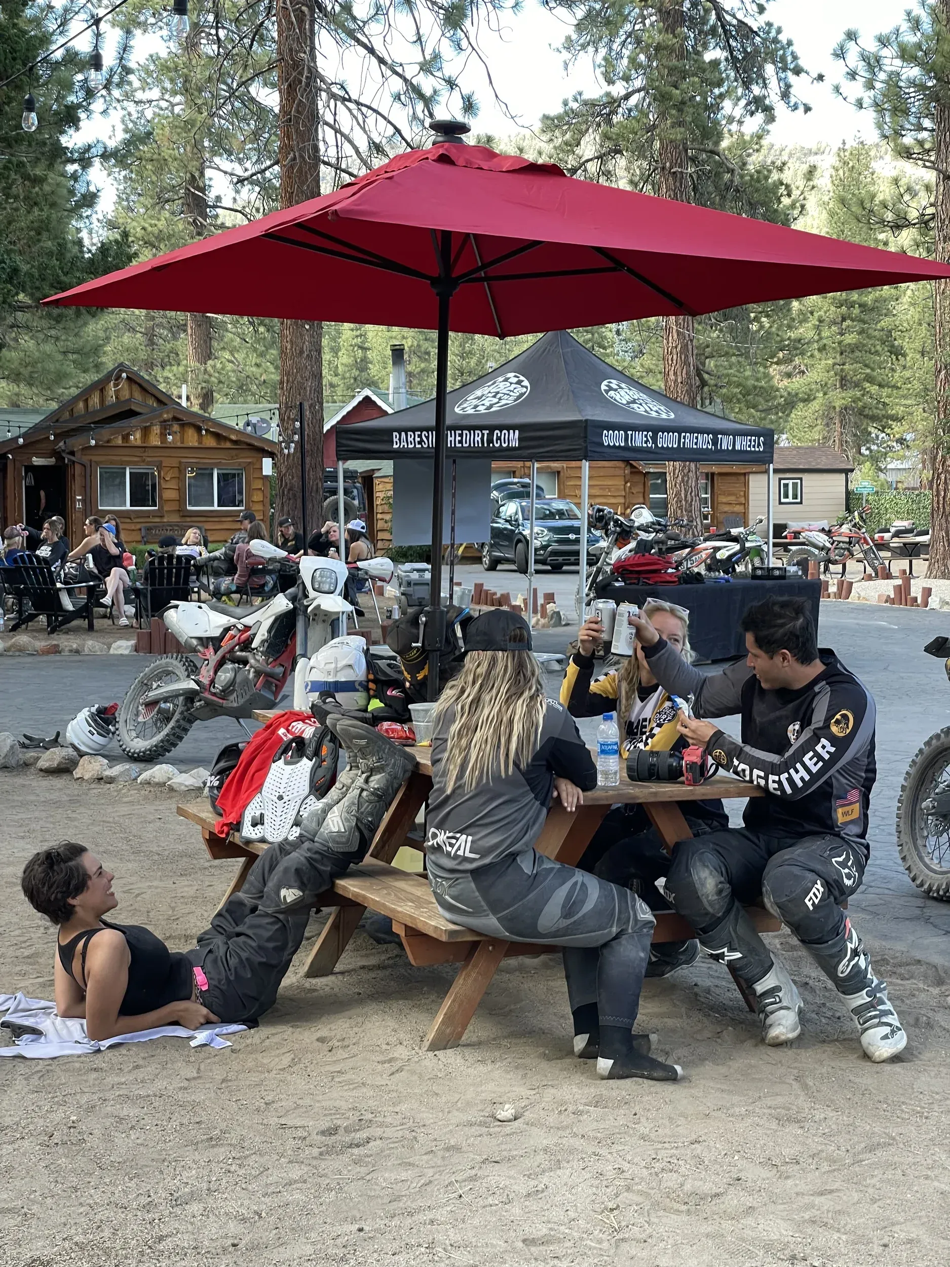 Group of people in motorcycle gear at picnic table under red umbrella.