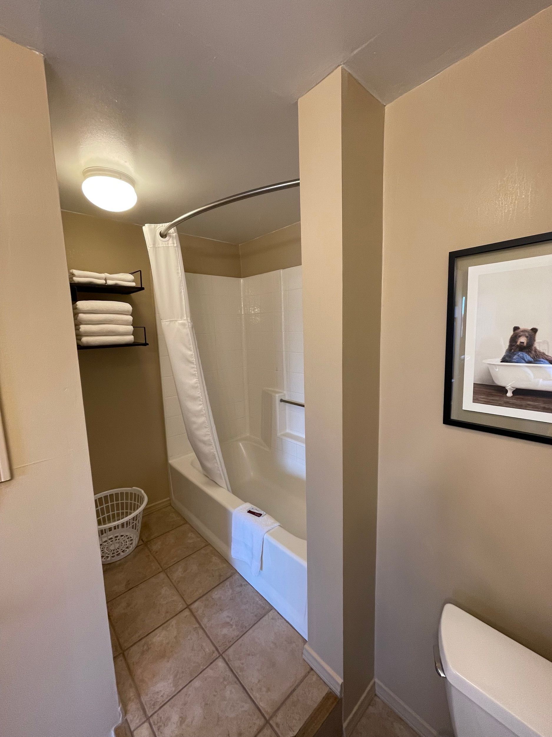Bathroom with a white bathtub/shower, towels on shelves, and a toilet. Beige walls.