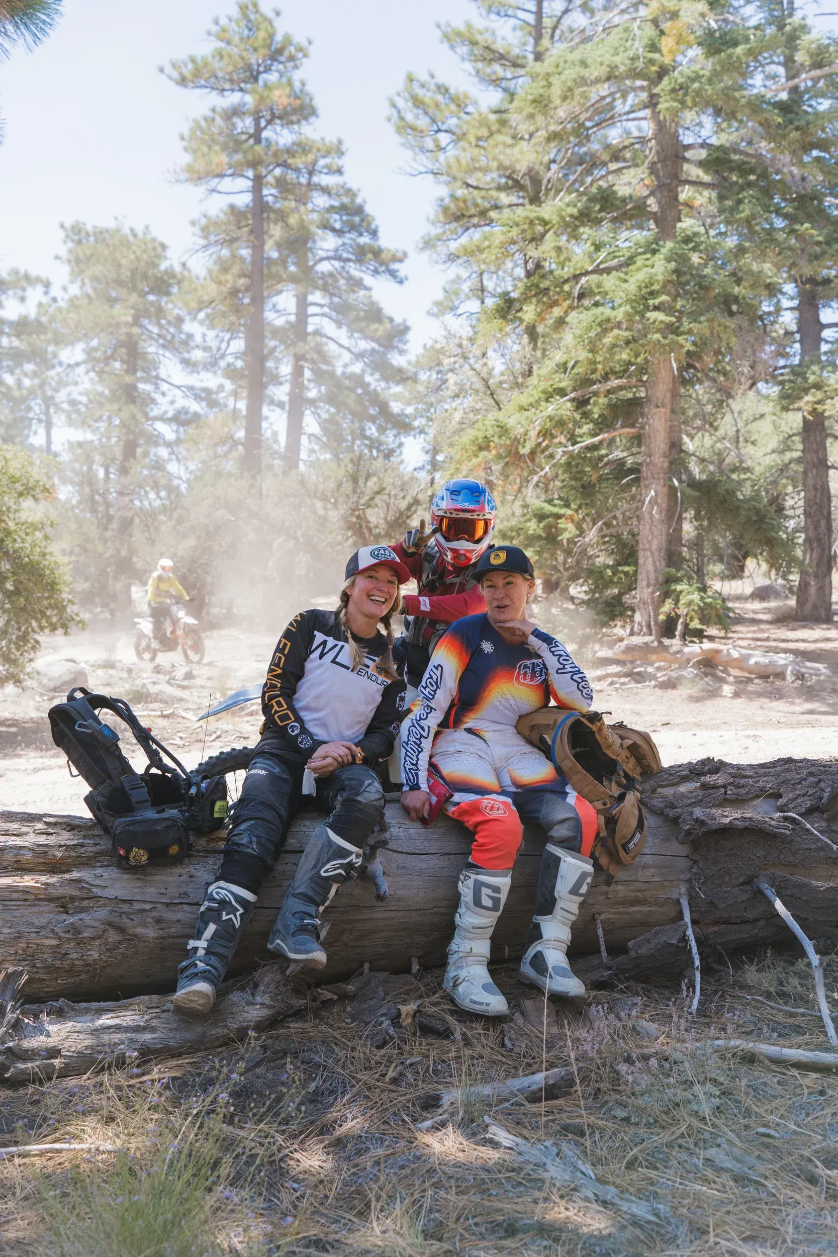 Three dirt bike riders sitting on a log in a forest, smiling.