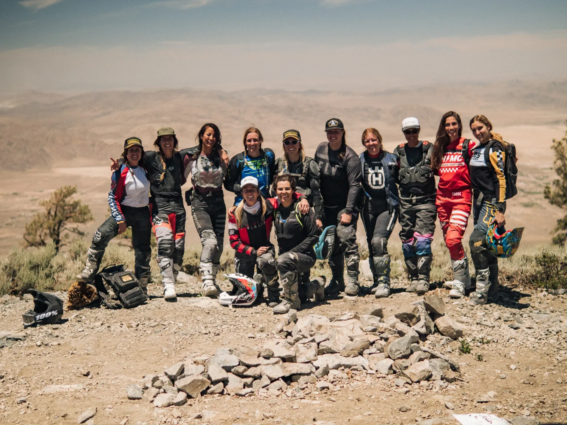 Group of dirt bike riders posing on a mountain, wearing helmets and gear, with a desert vista in the background.