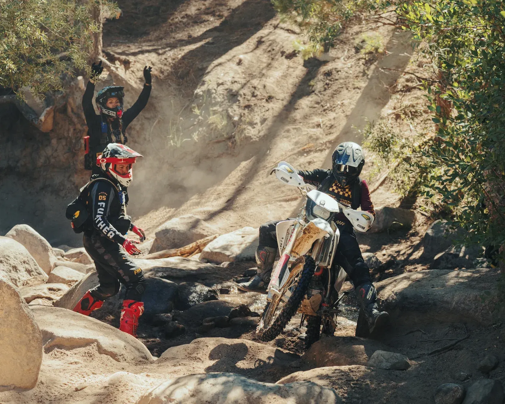 Three dirt bikers on a rocky trail, one on bike, two cheering. Dust and sunlight.