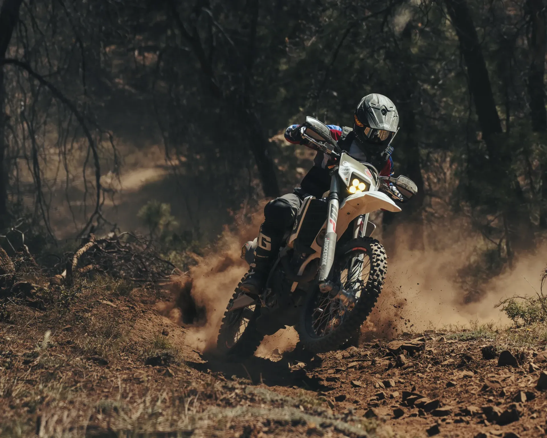 Dirt bike rider speeding on a dirt trail, kicking up dust; trees in background.