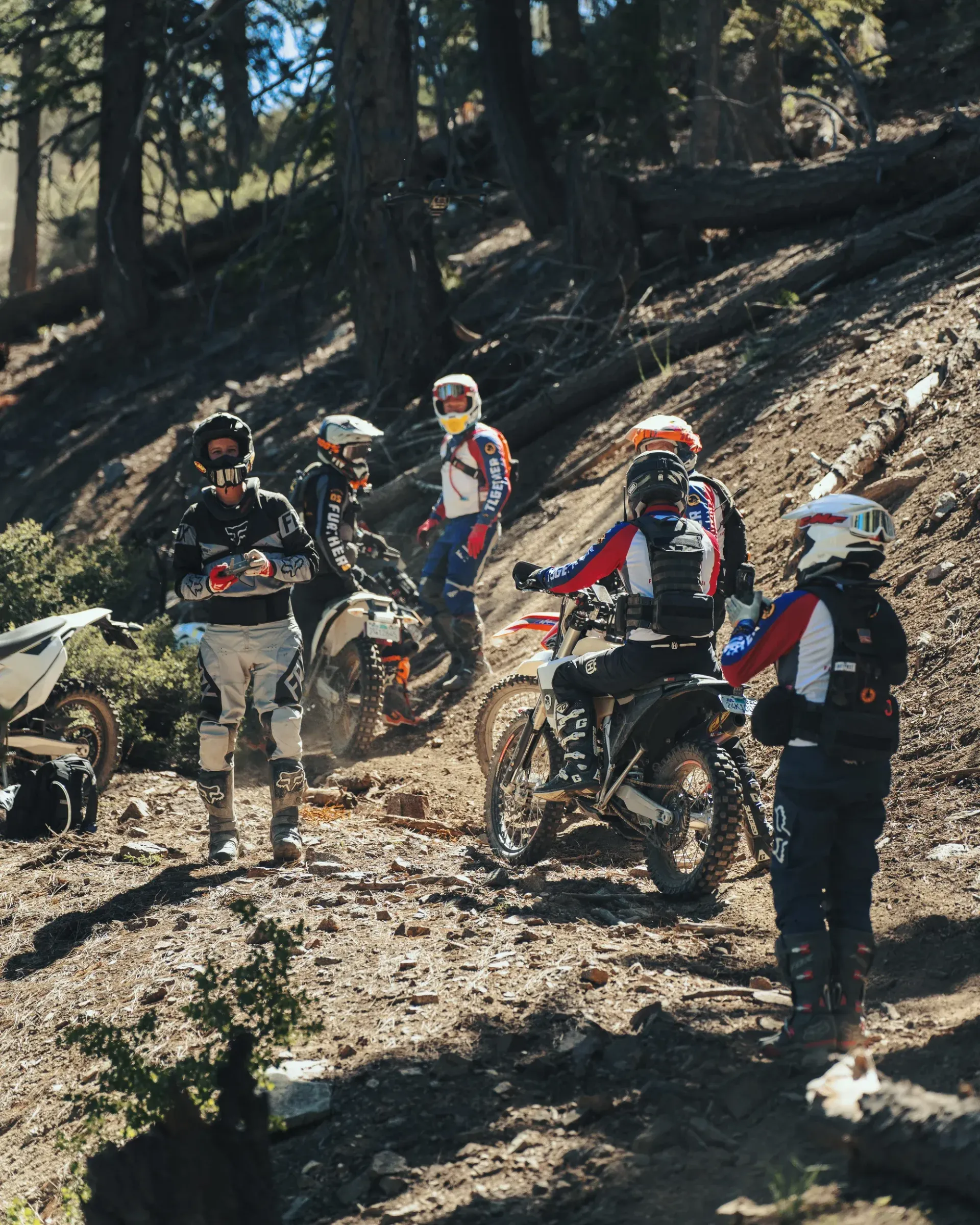 Group of dirt bikers on a trail in a forest, some standing, some on bikes.