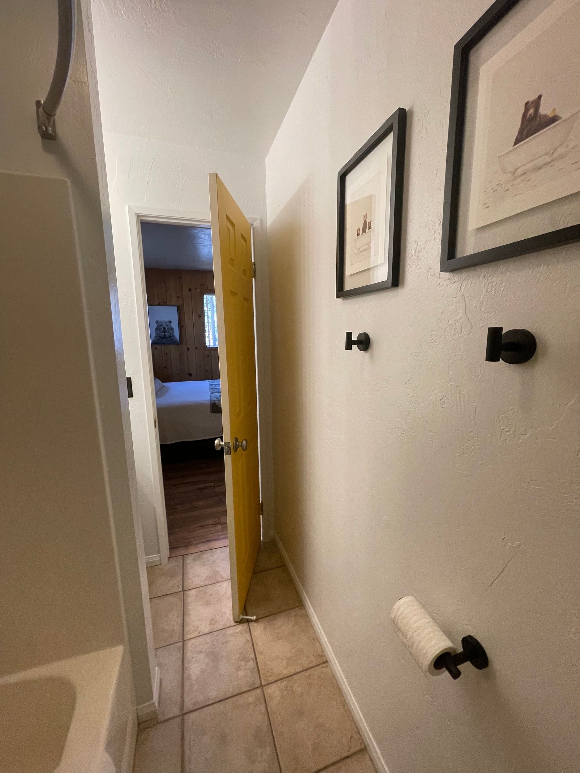 Narrow hallway with open yellow door to bedroom; tile floor, black fixtures on wall.