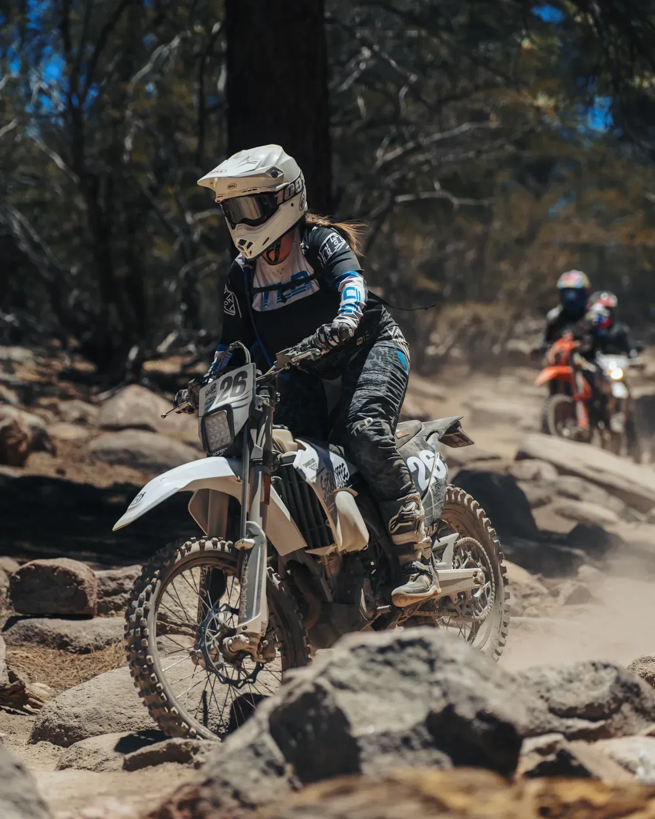 Woman on white dirt bike riding on a rocky trail with two riders behind.