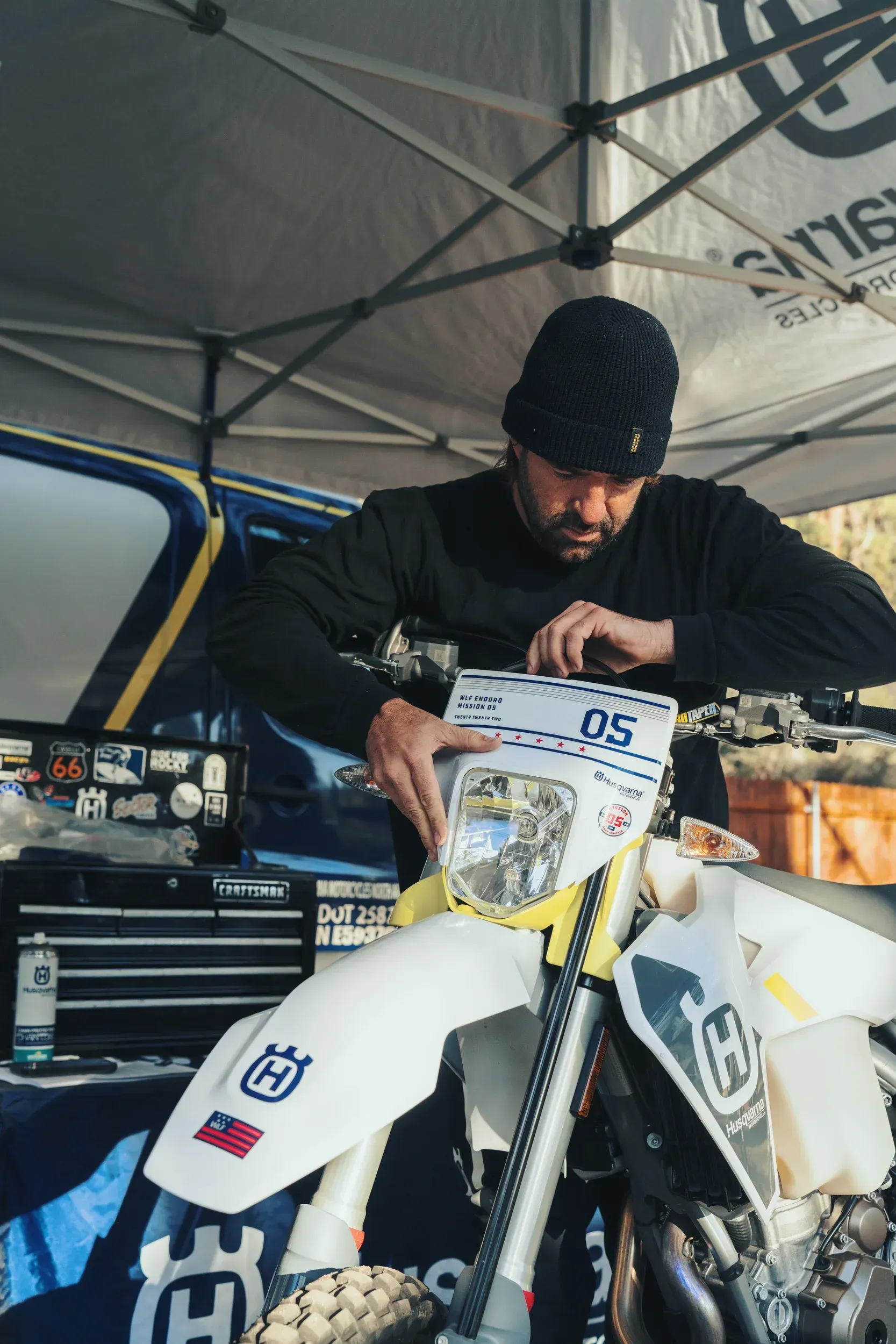 Man in black beanie works on a white Husqvarna motorcycle under a tent.
