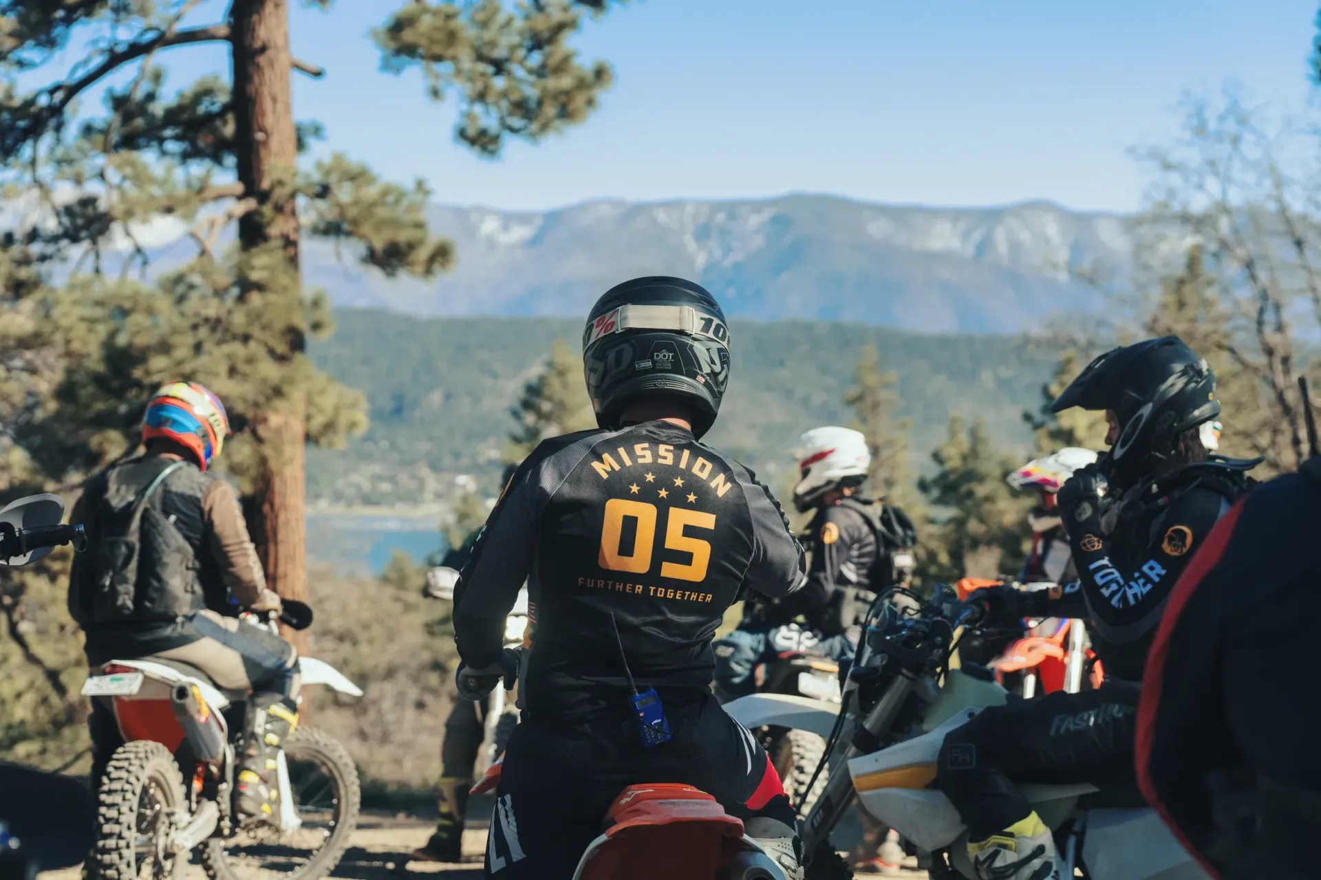 Motorcyclists in riding gear on bikes, overlooking a lake and mountains. The rider closest has 