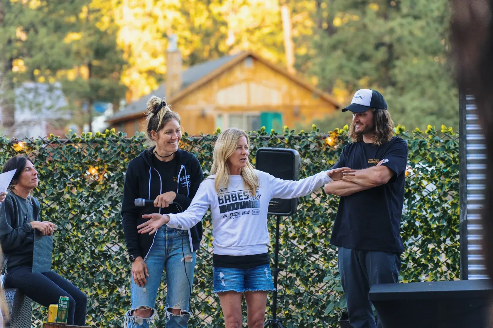 Four people on a stage outside, in front of greenery and a house. Two women and a man are gesturing.