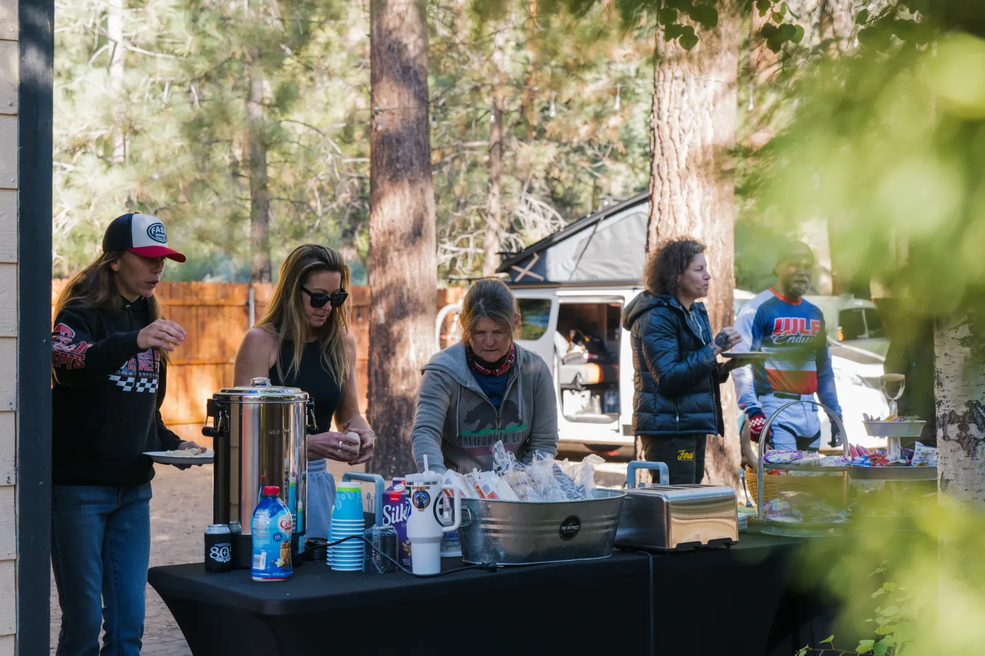 People at an outdoor food table with coffee, cups, and snacks. Trees and a van are in the background.