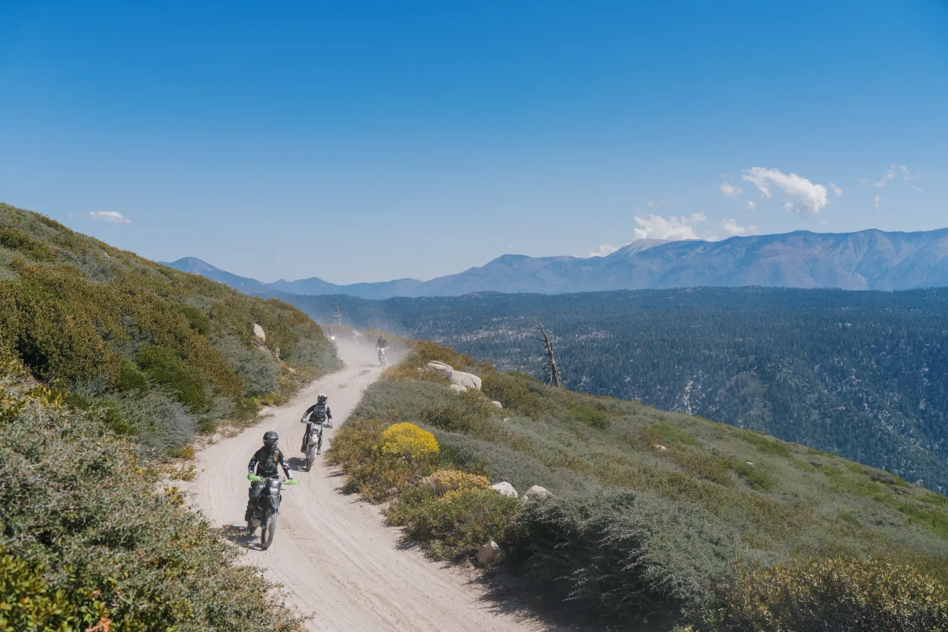Motorcyclists ride along a dirt road on a mountainside under a bright blue sky.