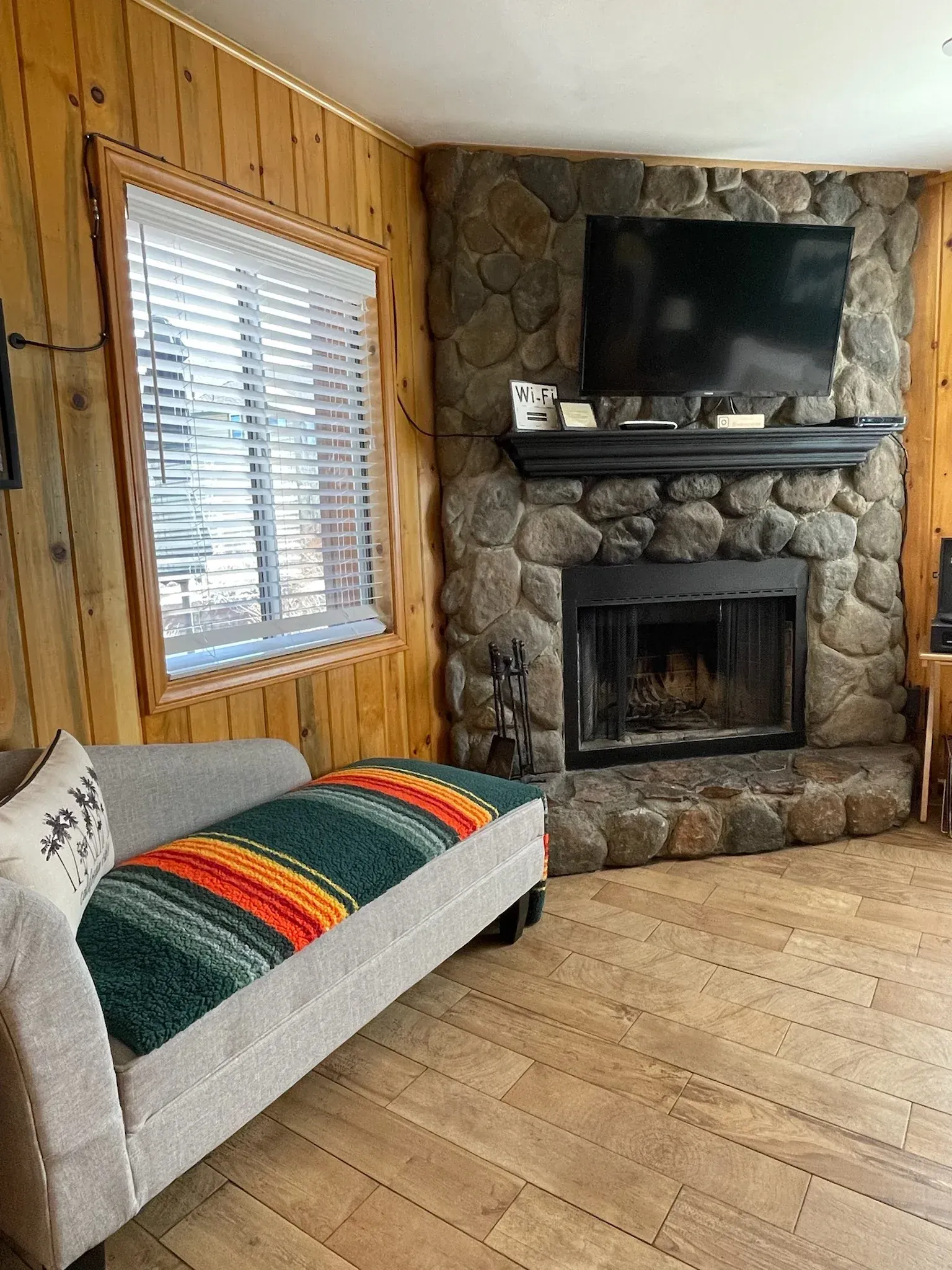 Living room with a stone fireplace, TV, couch, window with blinds, and wood paneling.