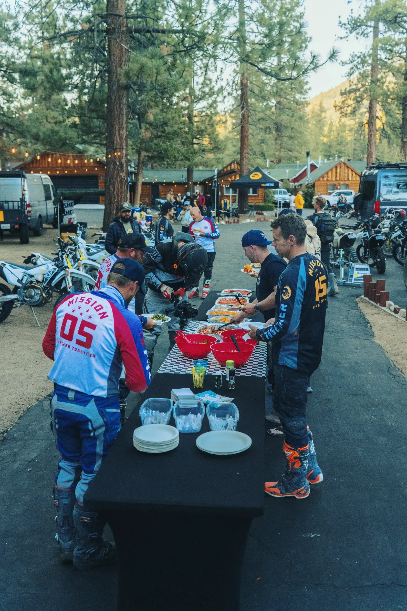People in motocross gear at a buffet table outdoors, eating. Pine trees and buildings in the background.
